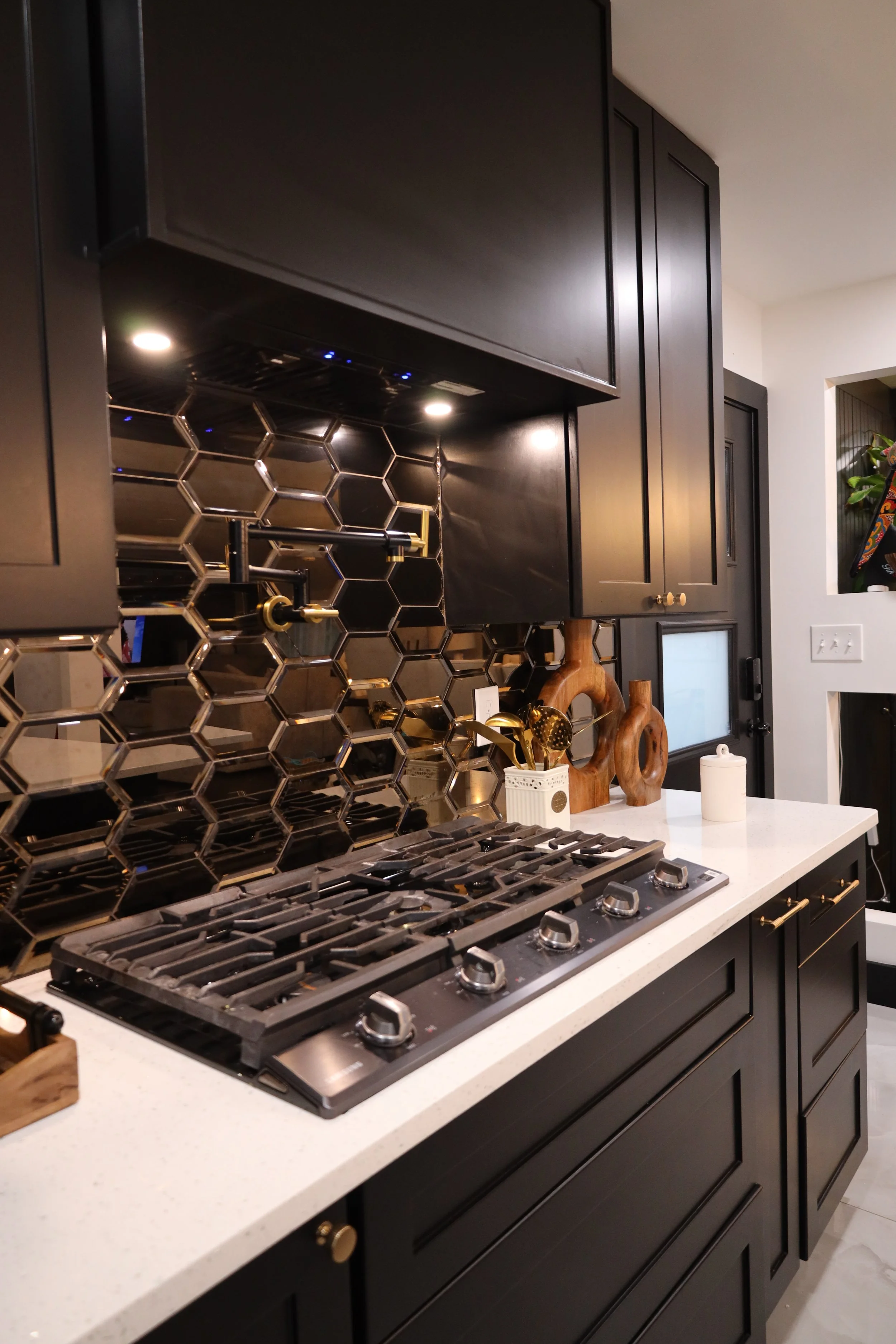Modern kitchen with black cabinets, white speckled countertop, and gold hardware. Black hexagonal tile backsplash with reflective surfaces and gold accents. Gas stove with six burners, wooden decor, and a white container on the countertop.