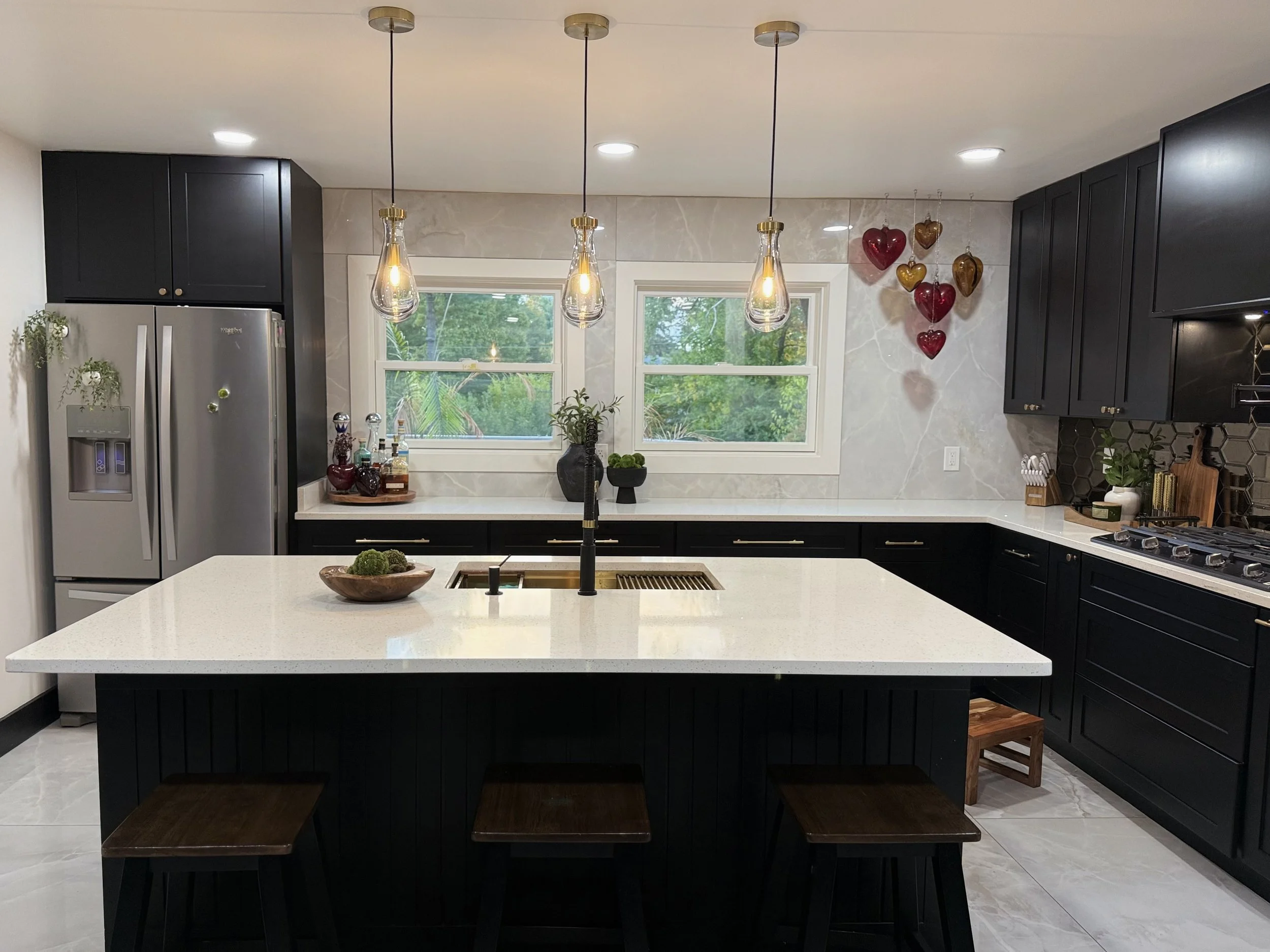 Modern kitchen with black cabinets, white countertop and island, stainless steel refrigerator, hanging pendant lights, and decorative hearts on the wall.