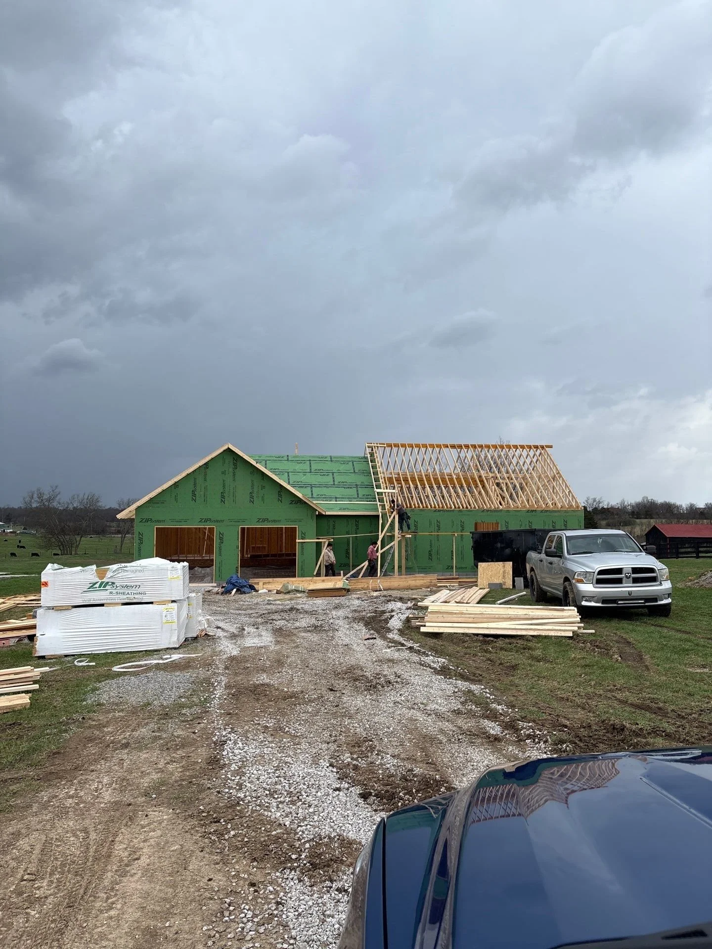 House under construction with a green exterior and wooden framing on the roof. Construction workers are working on the roof. A silver pickup truck is parked nearby, and construction materials are scattered on the gravel driveway. A dark stormy sky is in the background.