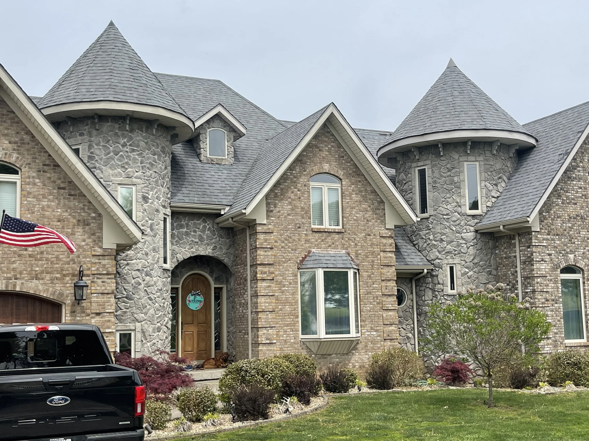 Large brick and stone house with turrets, multiple windows, and a wooden front door. A black Ford pickup truck is parked in front, and an American flag is mounted near the entrance. There are landscaped bushes and a small tree in the front yard.