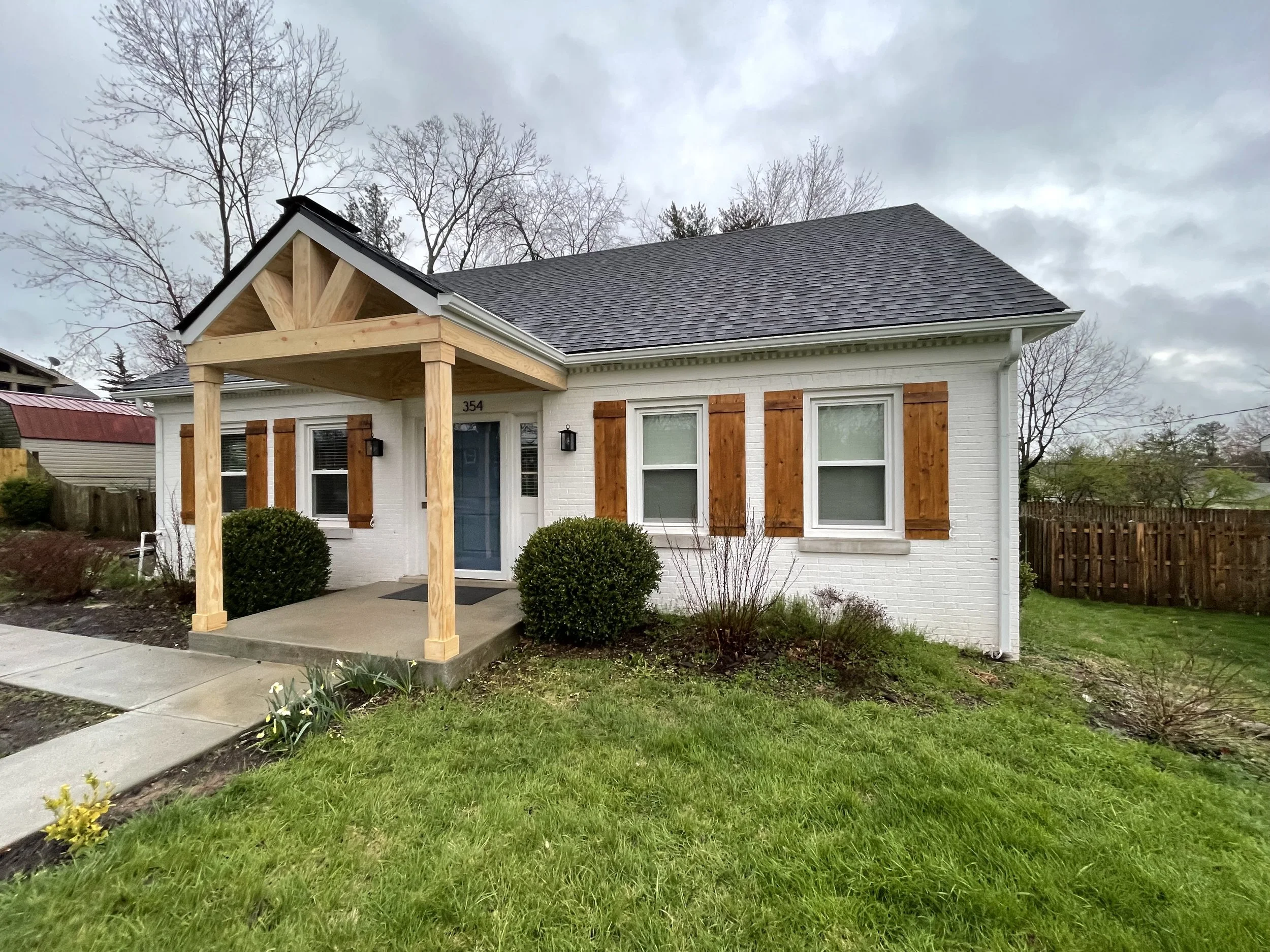 Exterior of a small white brick house with a porch supported by four wooden posts, brown shutters, and a gray shingled roof. Green bushes are in front, and a sidewalk leads to the porch. The yard has grass and some plants, and the sky is cloudy.