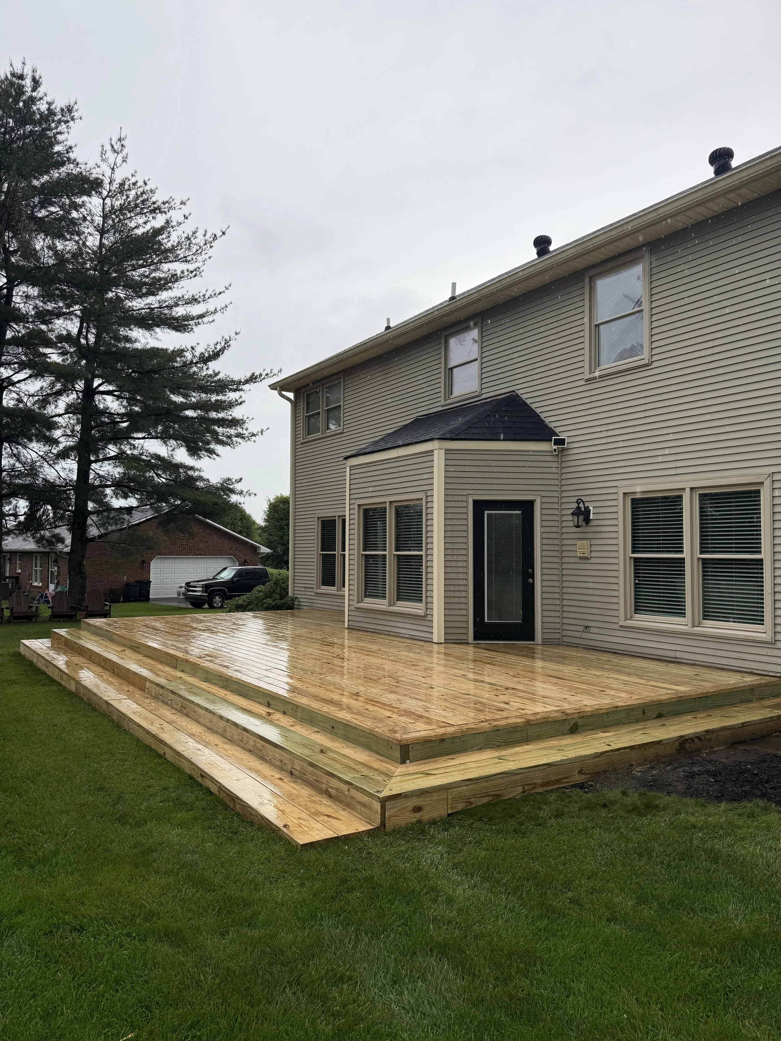Backyard with a newly built wooden deck attached to a beige house with multiple windows, on a rainy day.