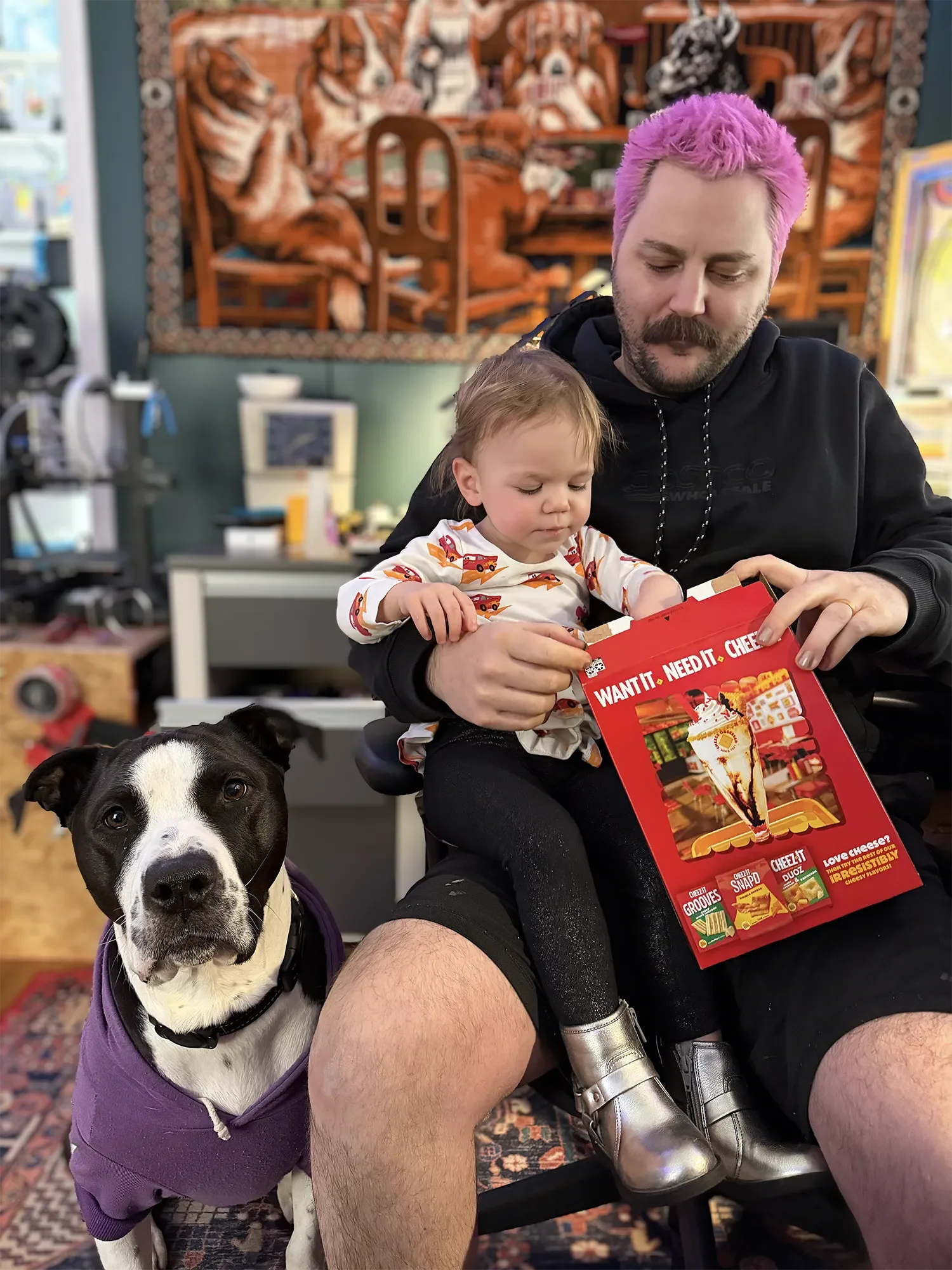 A man with pink hair sitting with a young child and a black and white dog. The man and child are examining a red box of Cheez-It snack crackers. The background has colorful artwork and shelves with various items.