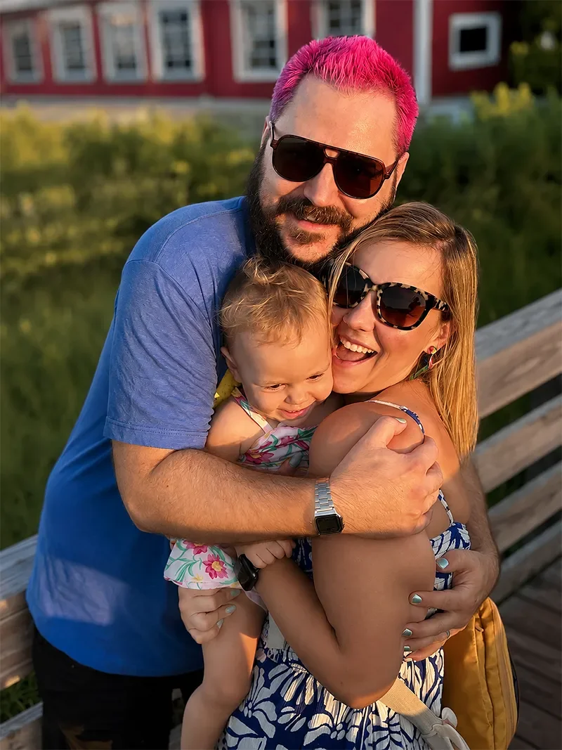 A happy family of three hugging outdoors on a wooden bridge during sunset, all wearing sunglasses and smiling.
