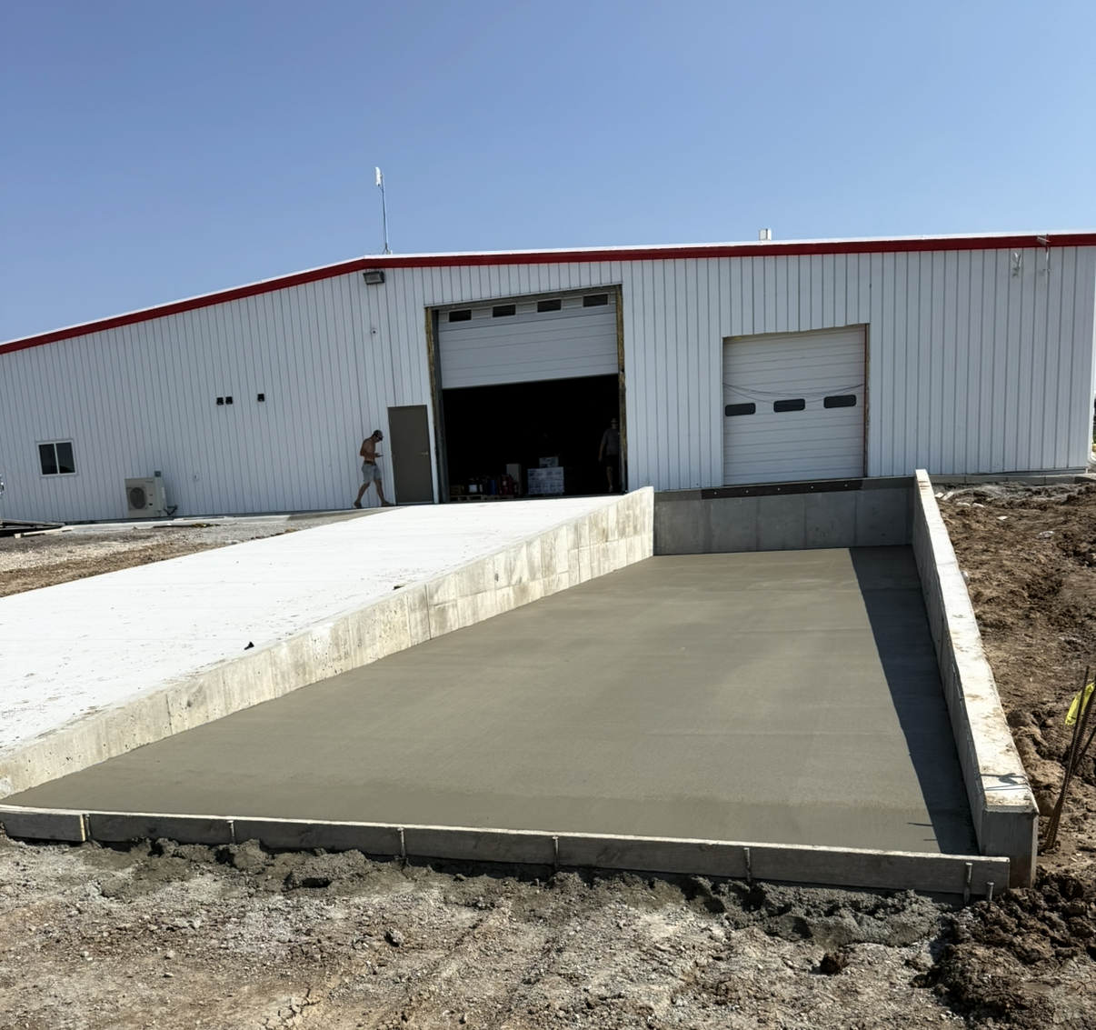 Newly poured concrete slab in front of a warehouse with large roll-up doors, under a clear blue sky.