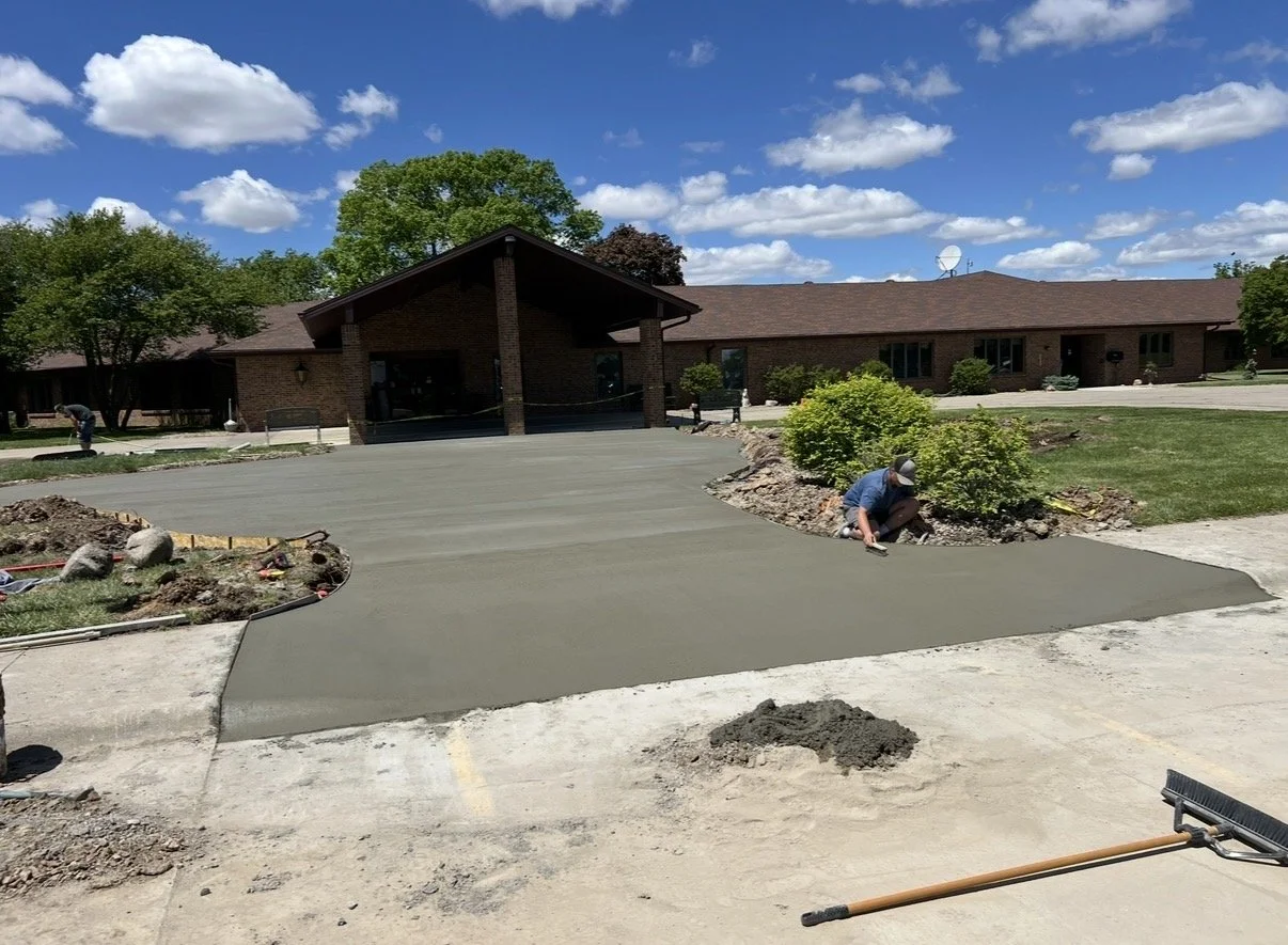 Workers are installing fresh concrete on a driveway in front of a brick building under a partly cloudy sky.