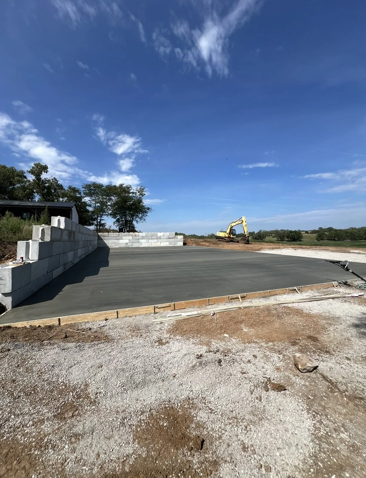 Construction site with a freshly poured concrete slab, a small yellow excavator in the background, and a partially built cinder block wall to the left under a blue sky with scattered clouds.