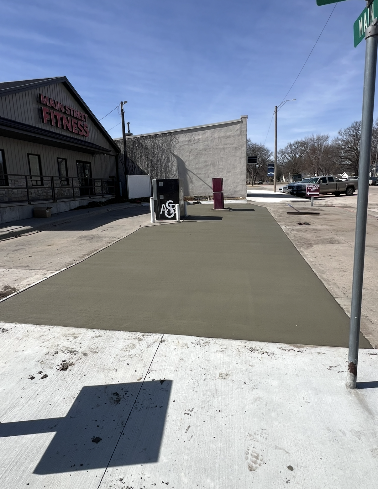 Newly poured concrete sidewalk in front of a fitness center with a sign for Main Street Fitness, and a black A and G on it. There are some footprints and dirt on the sidewalk.