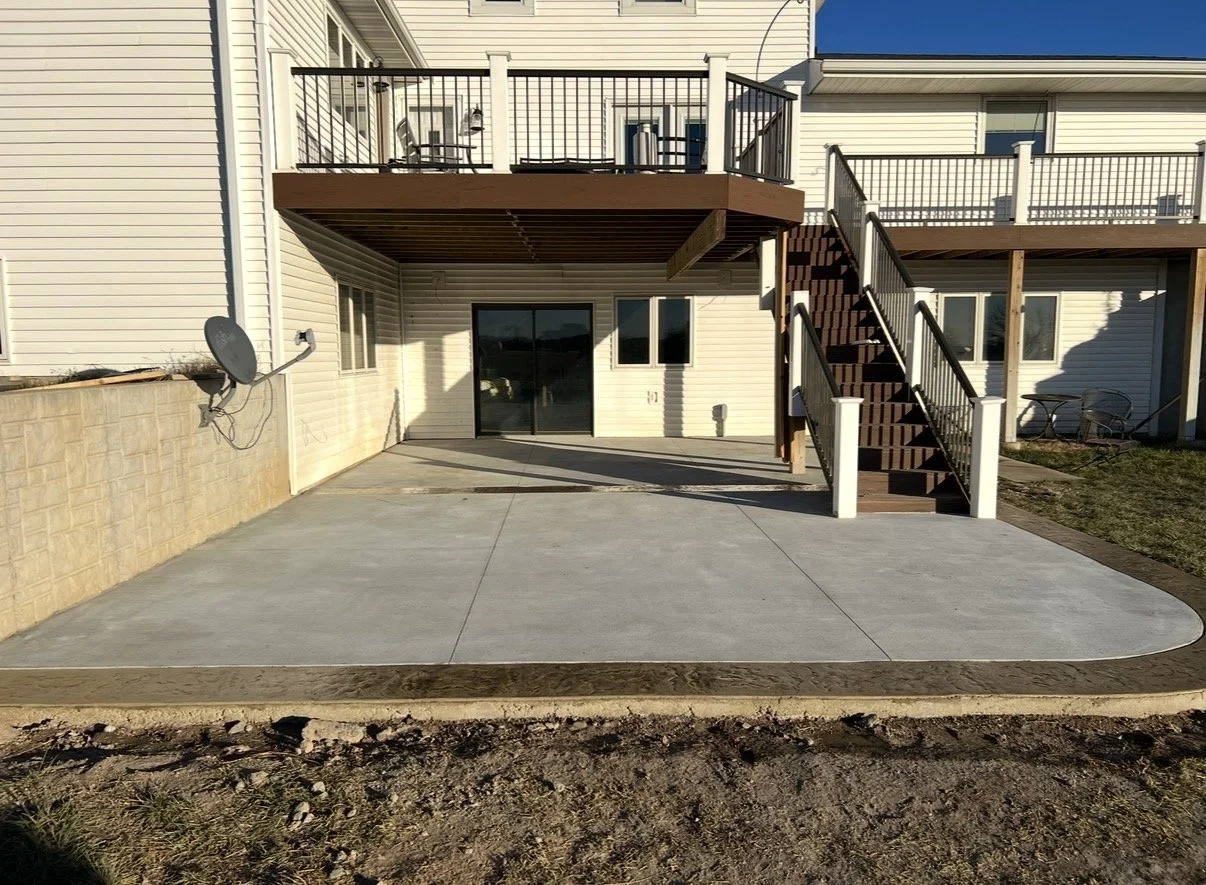 Backyard patio area with a concrete surface, a staircase leading to an upper deck, and a satellite dish attached to the beige house wall.