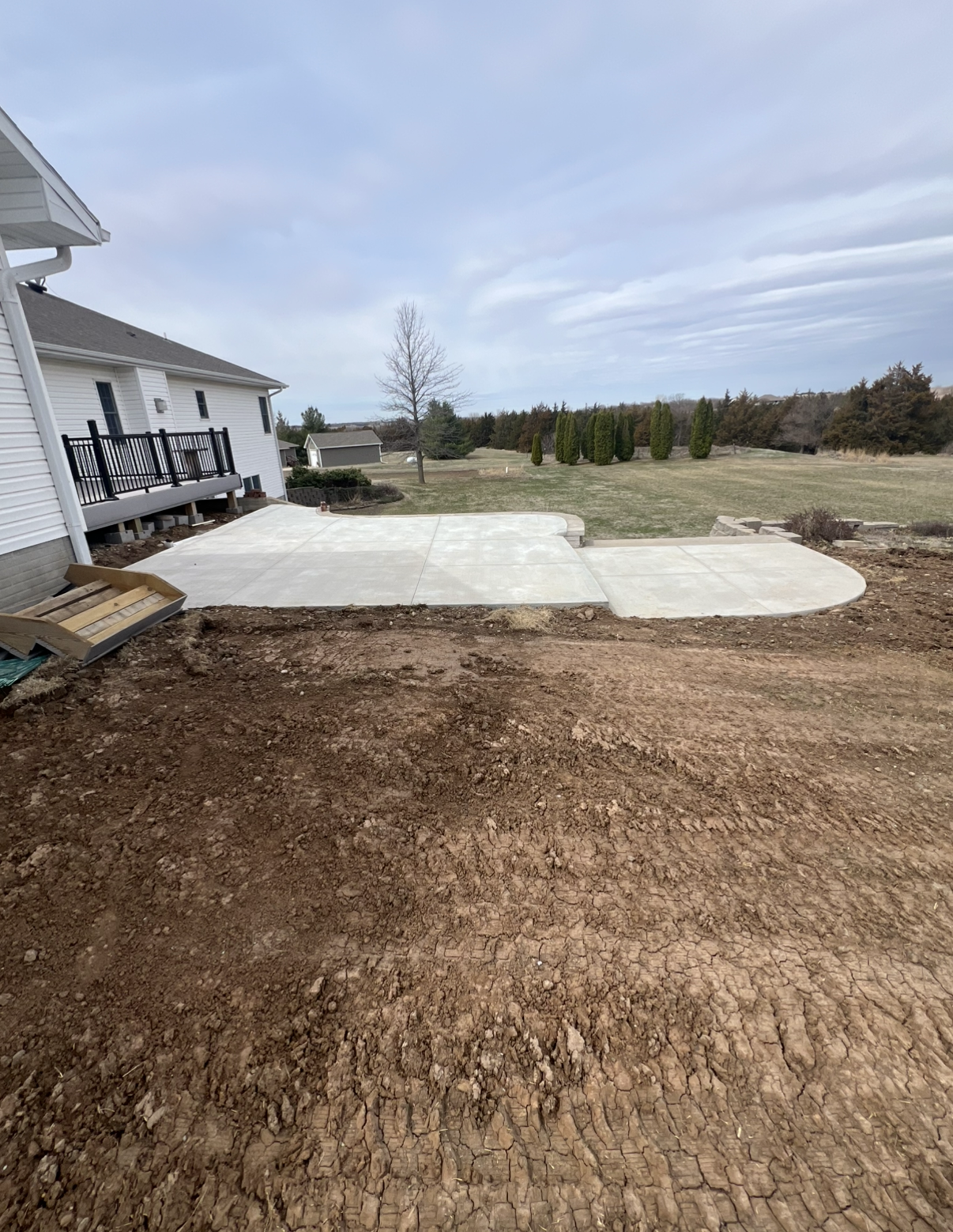 Newly poured concrete patio with a curved edge, adjacent to a house with a black railing deck, and a grassy backyard with trees in the distance.