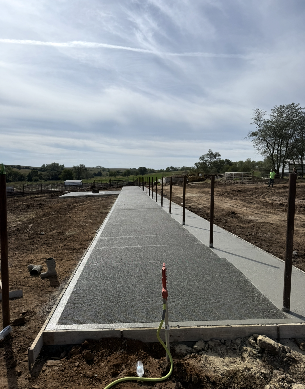 Newly constructed concrete pathway with metal railing posts in a rural outdoor setting, under a cloudy sky.