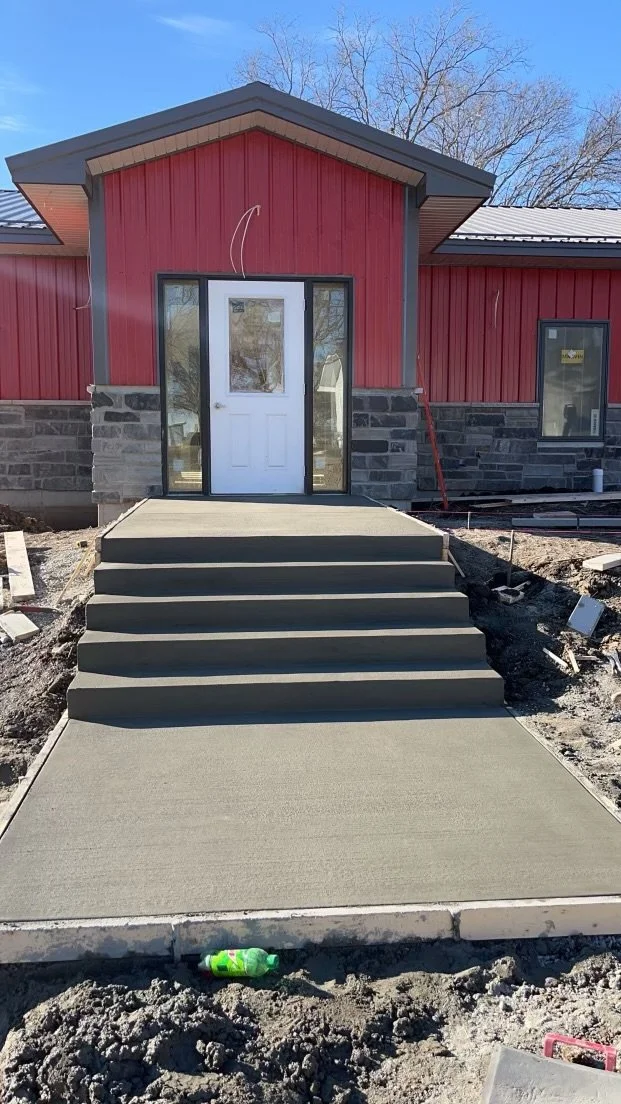Front entrance of a house with freshly poured concrete steps leading to a concrete porch, red exterior siding, and a white door with glass panels, under a cloudy blue sky.