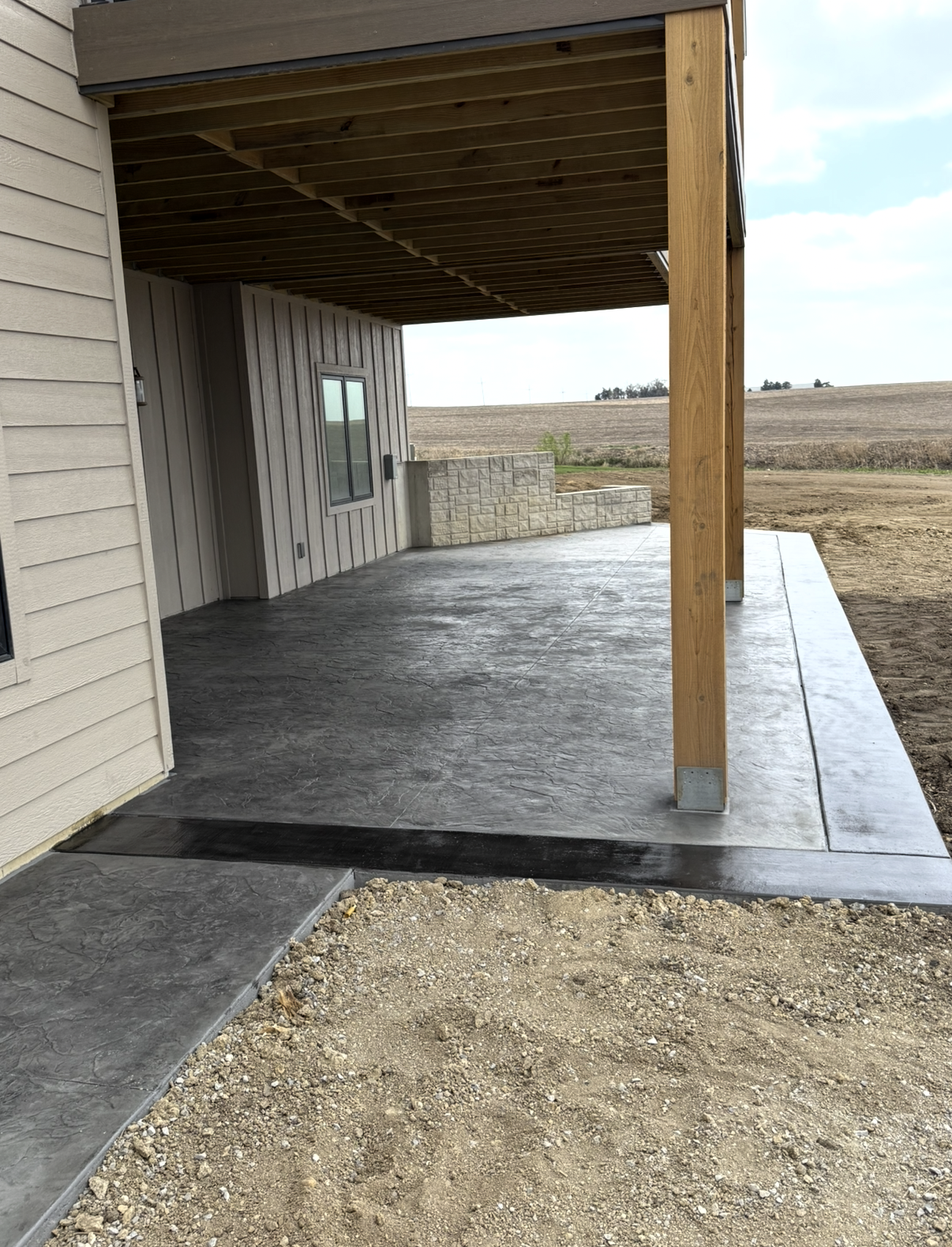 Concrete patio with textured finish under a wooden deck supported by wooden posts, adjacent to a house with beige siding and a small built-in brick wall in a rural area.