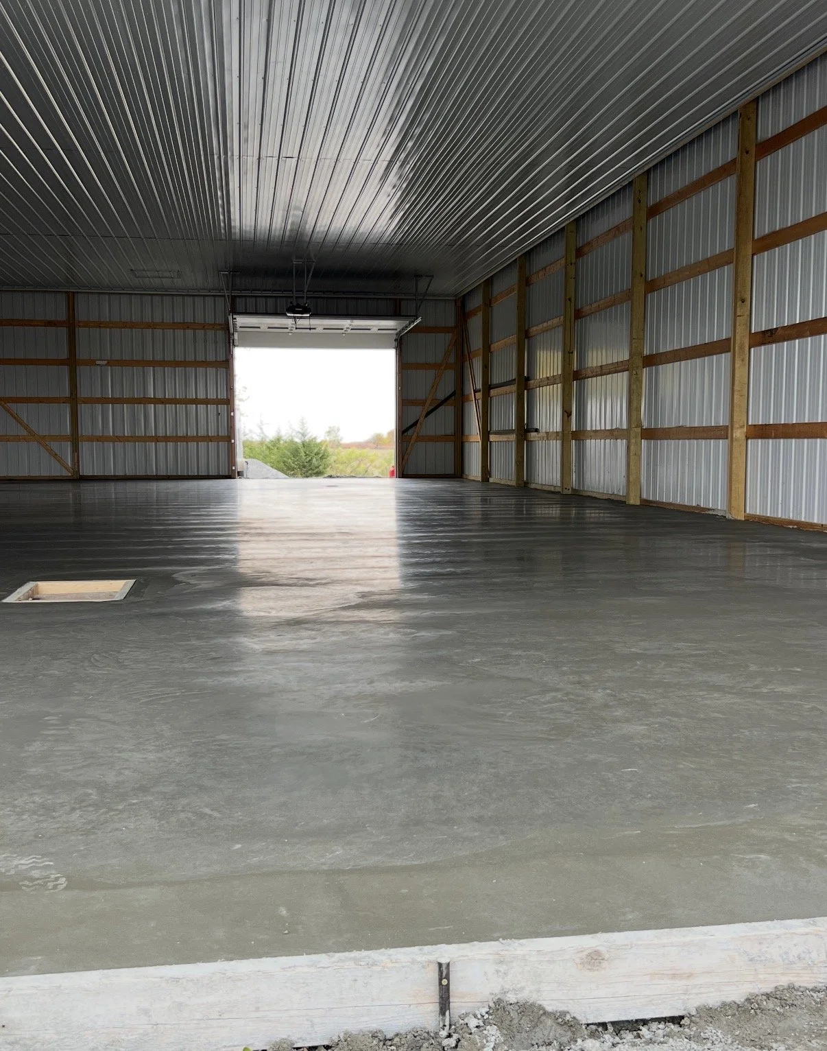 Empty industrial garage with a concrete floor, metal walls, and an open large garage door revealing trees outside.