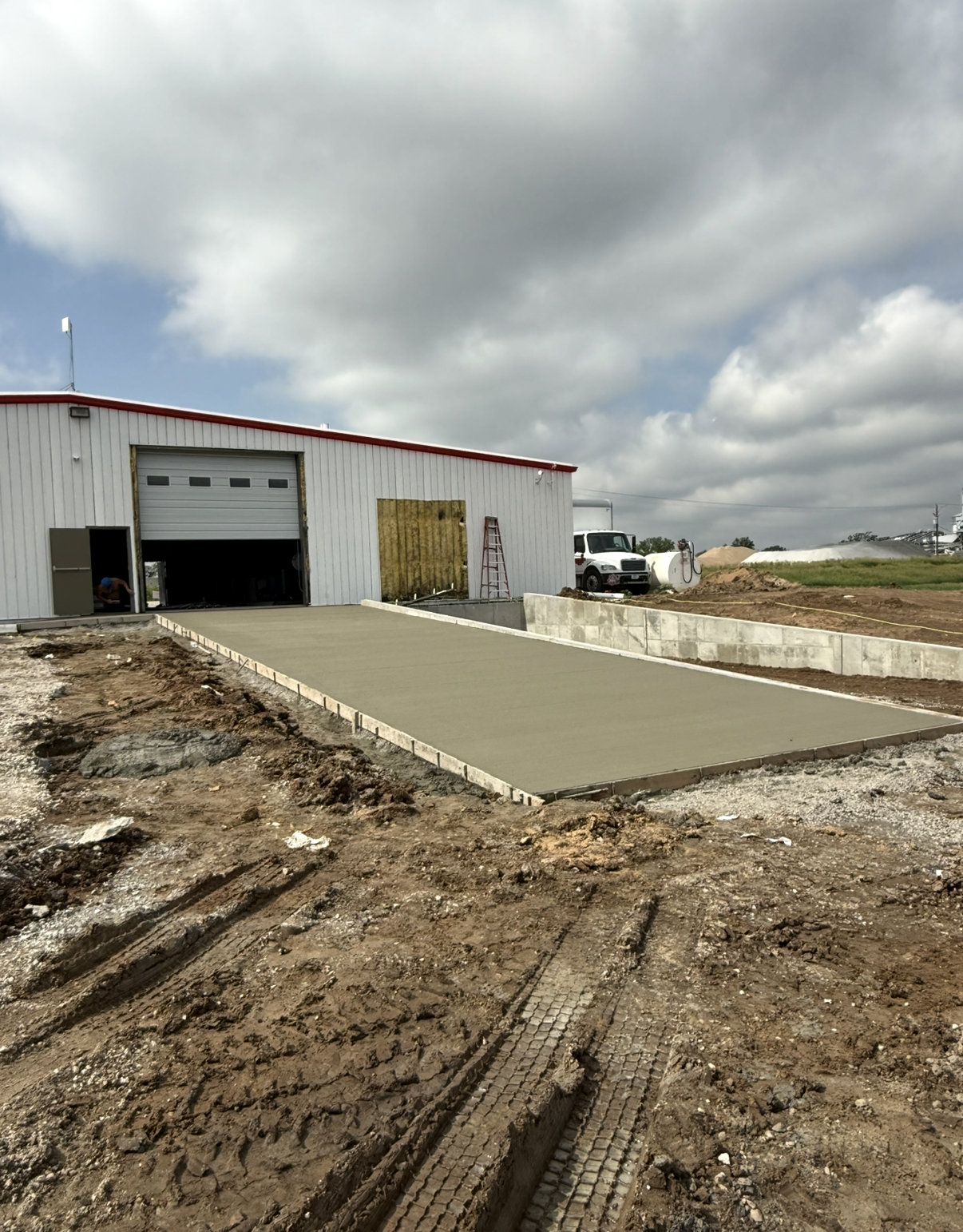 A concrete driveway is under construction in front of a warehouse with a partially open roll-up door, on a cloudy day.
