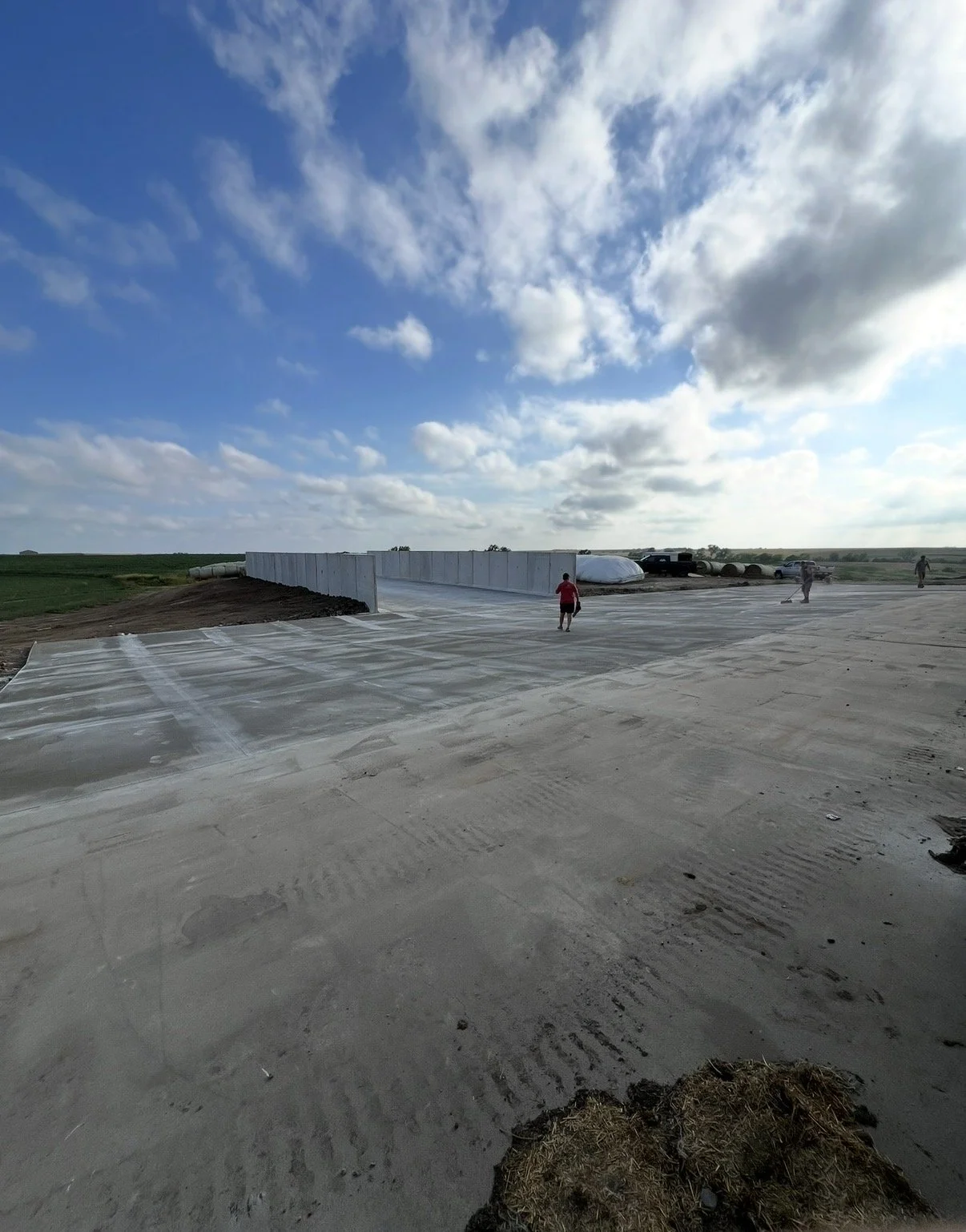 A construction site with a concrete slab foundation, a white wall in progress, and a partly cloudy sky overhead. Several workers are present.