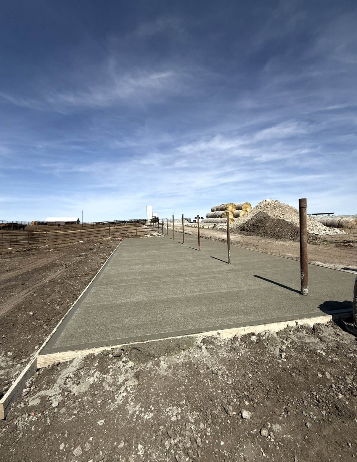 A newly poured concrete sidewalk under construction on a rural property with a clear blue sky and construction materials in the background.
