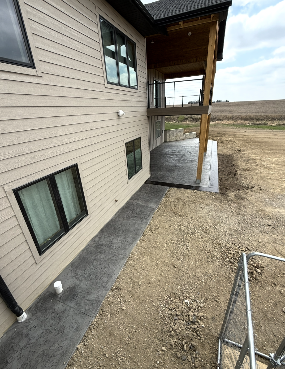 Exterior view of a new house with beige siding, black window frames, and a small concrete patio. There is a second-story balcony with a metal railing, supported by wooden posts. The surrounding area is mostly dirt and open land, with some constructio