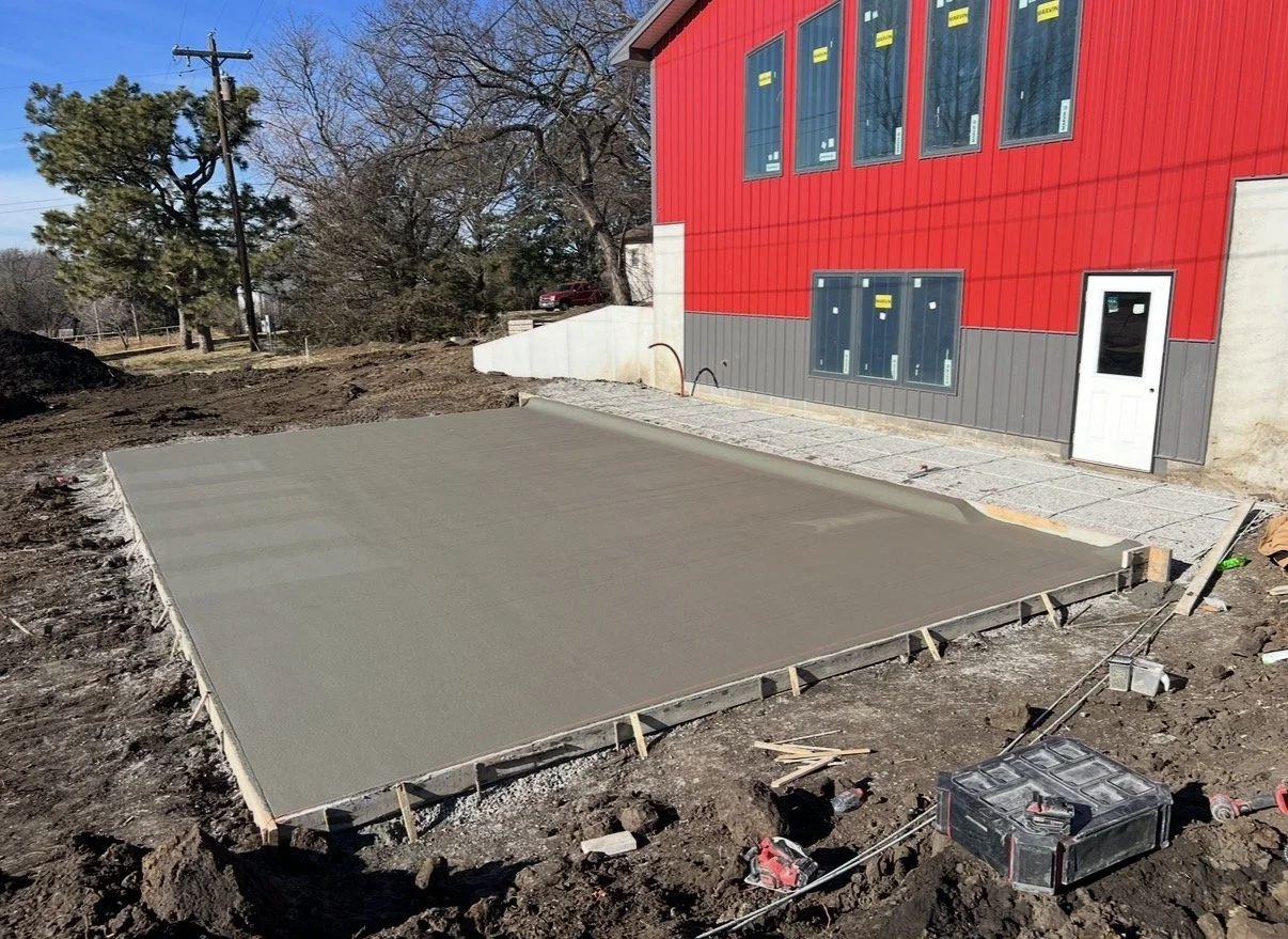 Concrete slab being poured in a backyard next to a red building with large windows and a door, with construction tools and materials nearby.