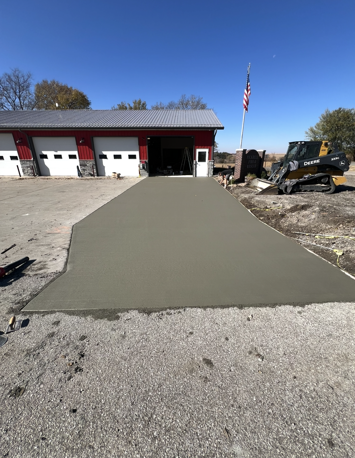 Concrete driveway being poured in front of fire station with hose and small construction equipment nearby, American flag flying on flagpole