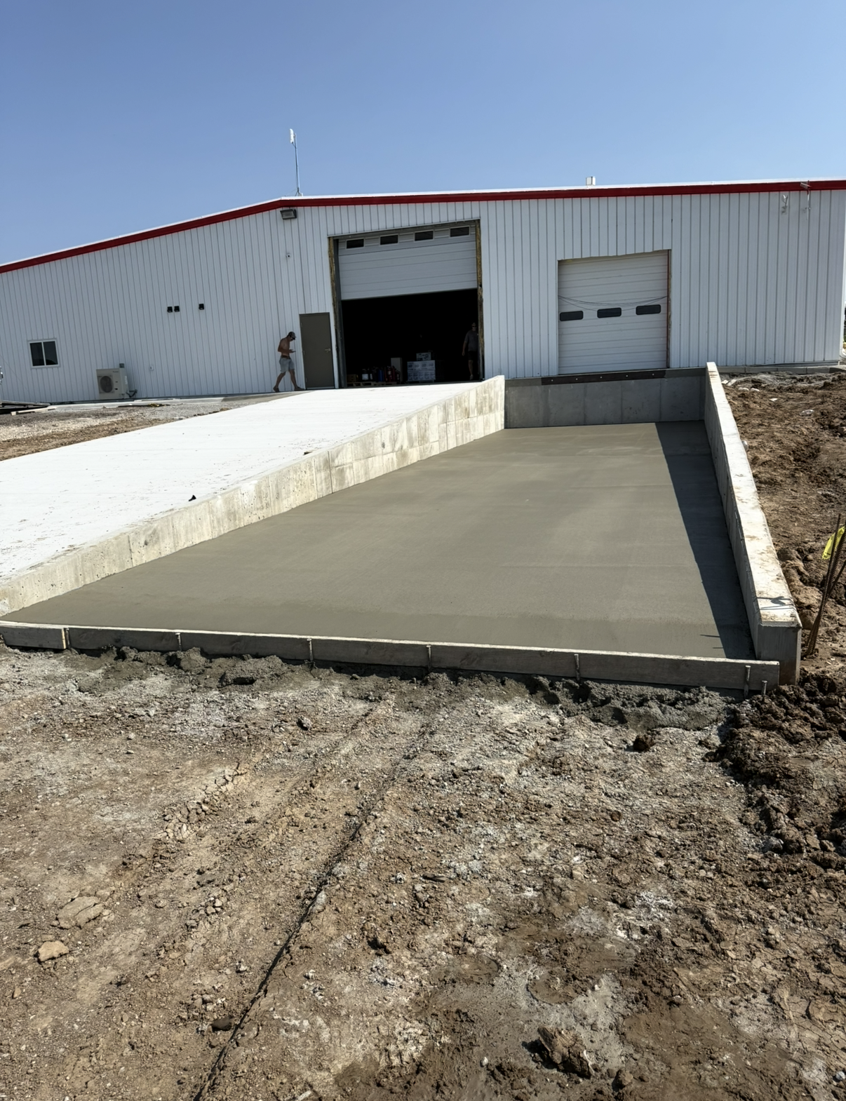 Freshly poured concrete concrete driveway leading to a large white metal building with open garage door andsmall window, surrounded by dirt and construction site.