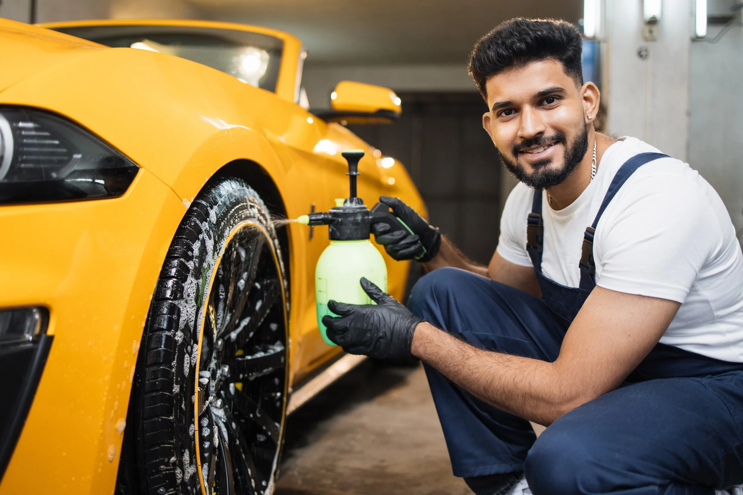 A man in a white t-shirt and overalls washing a yellow sports car in a garage, kneeling and holding a foam sprayer.