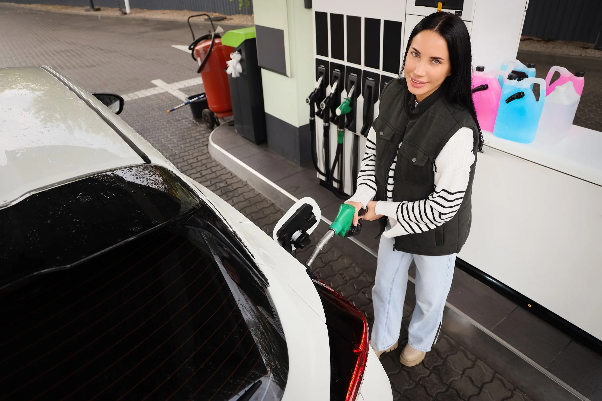 A woman washing her white and black car at a gas station with a pressure washer.