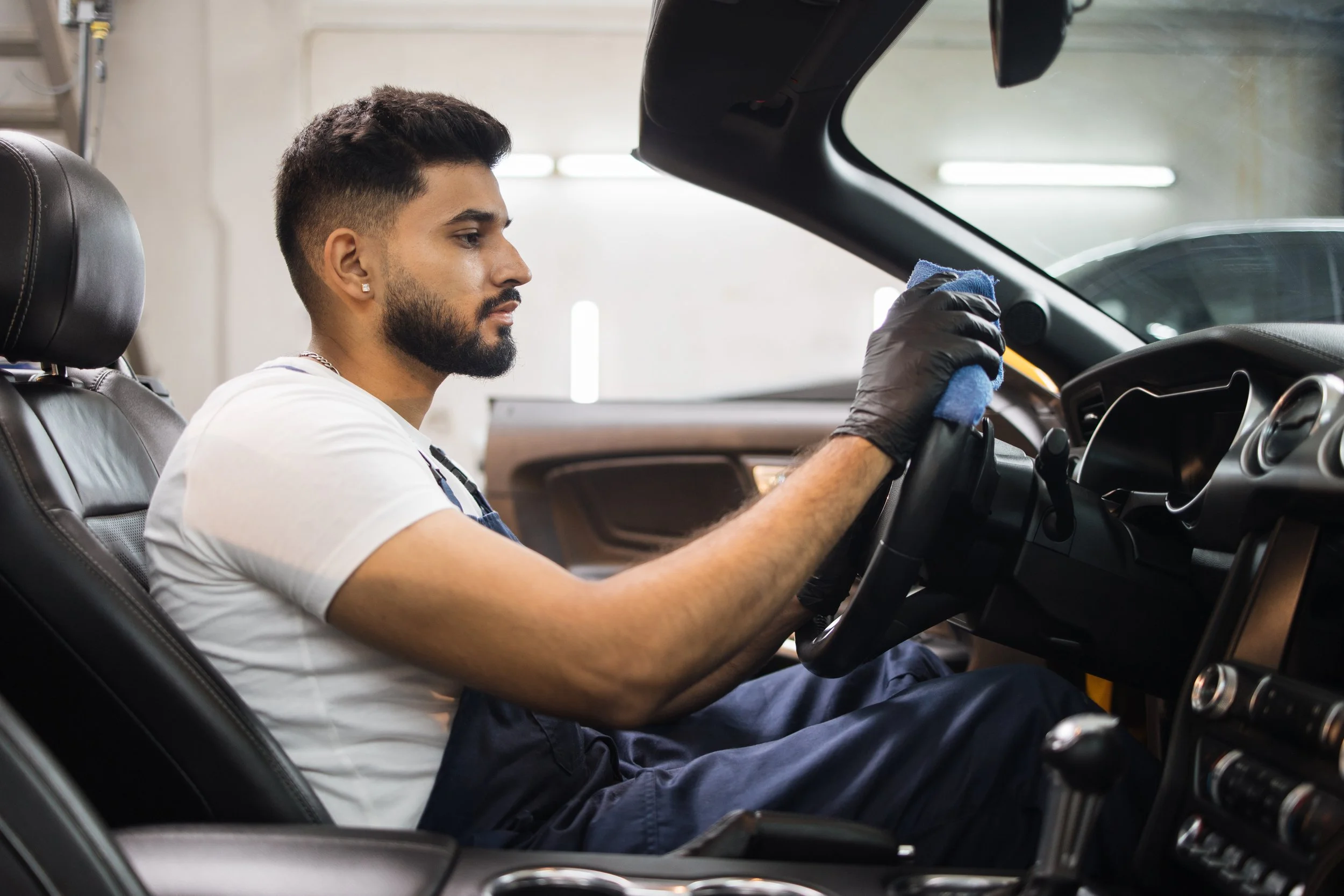 A man with short dark hair and a beard wearing a white t-shirt and black gloves cleaning the interior of a car, focusing on the steering wheel.