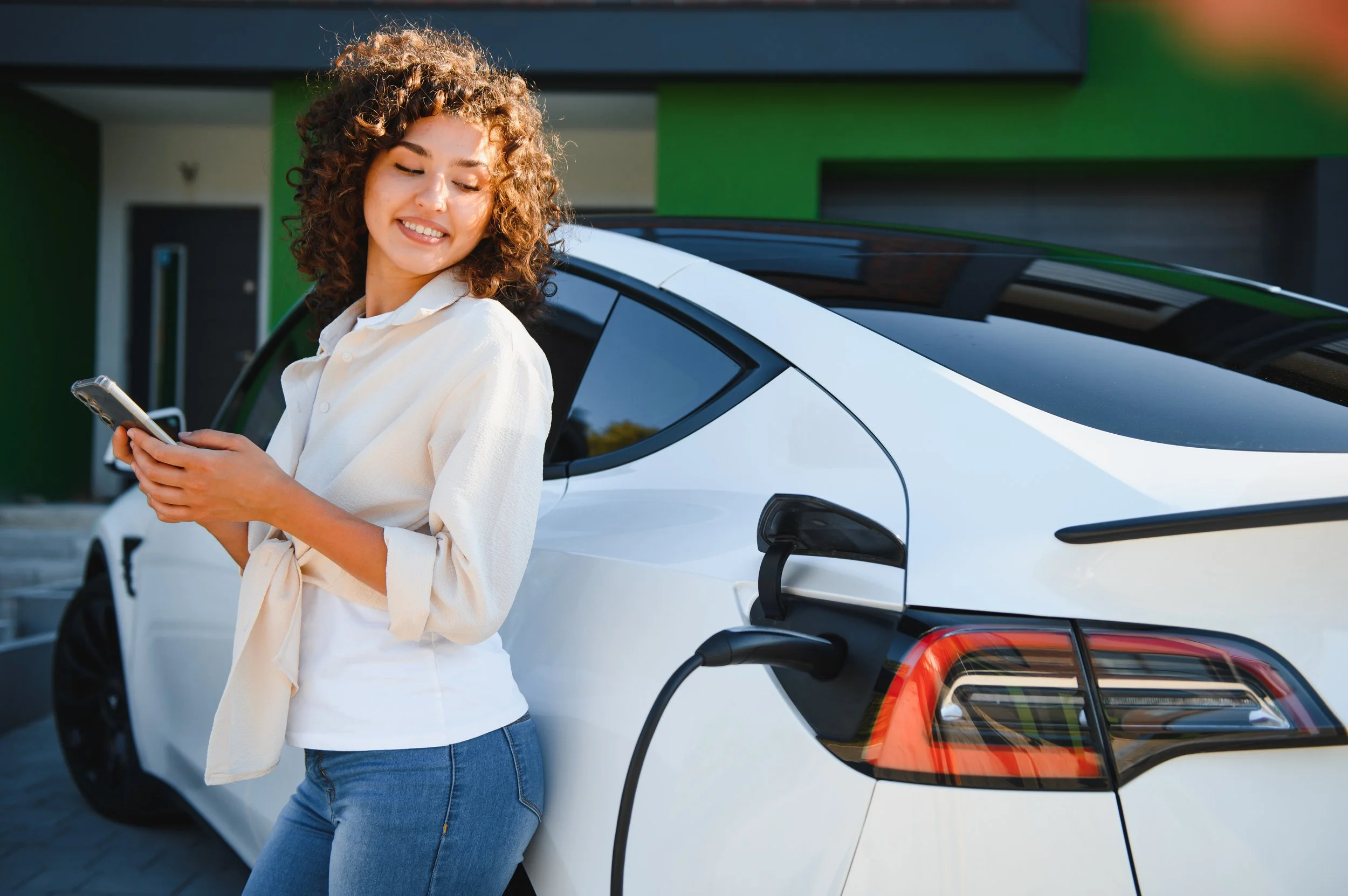 A woman with curly hair smiling and looking at her phone while standing next to an electric car that is plugged in for charging.