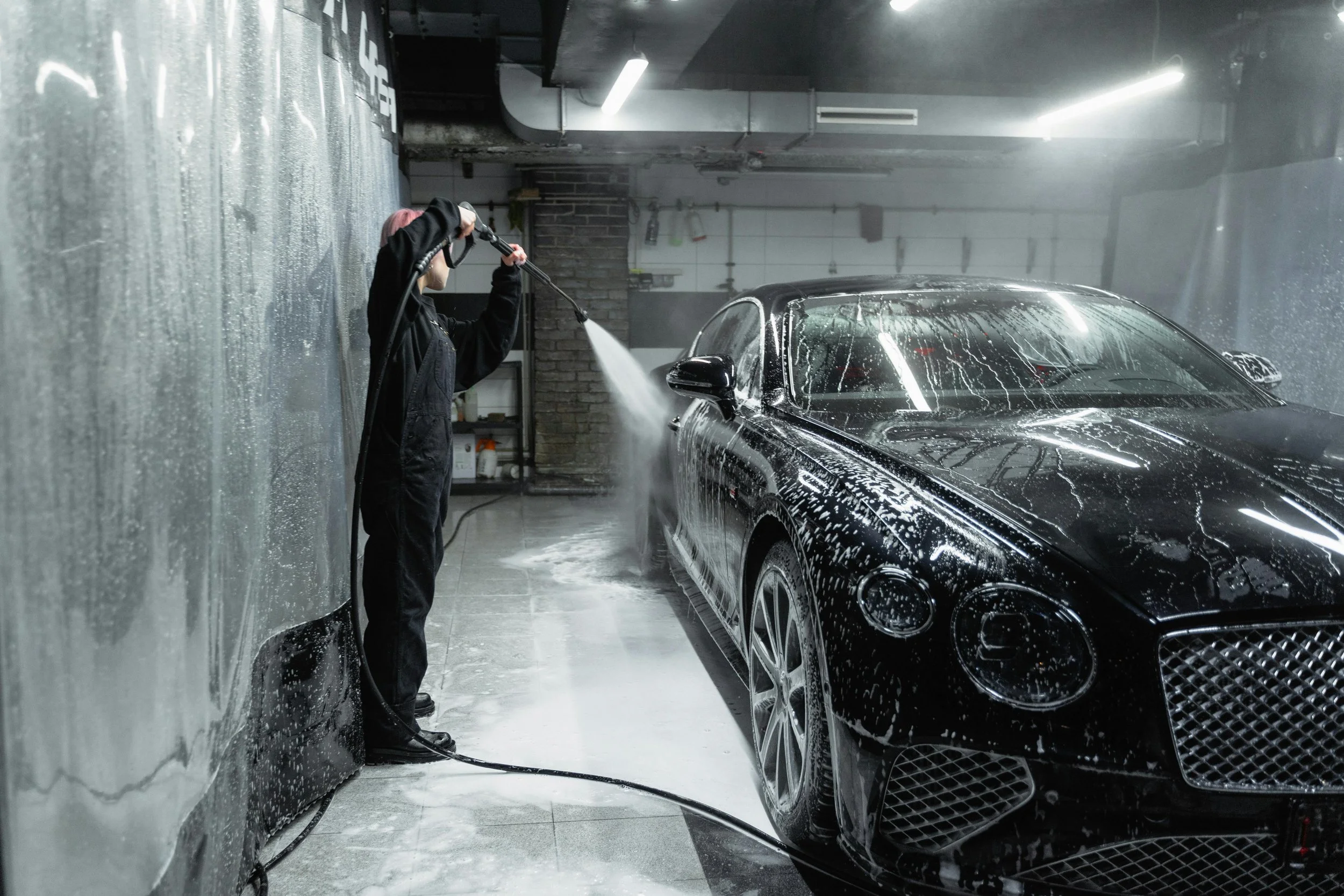 Person washing a black luxury car with a hose inside an industrial-style garage.