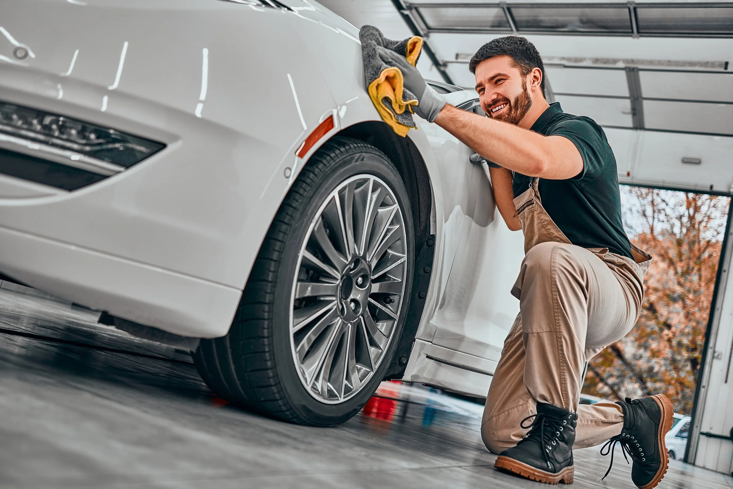 A man kneeling next to a white car in a garage, working on the wheel with a cloth.