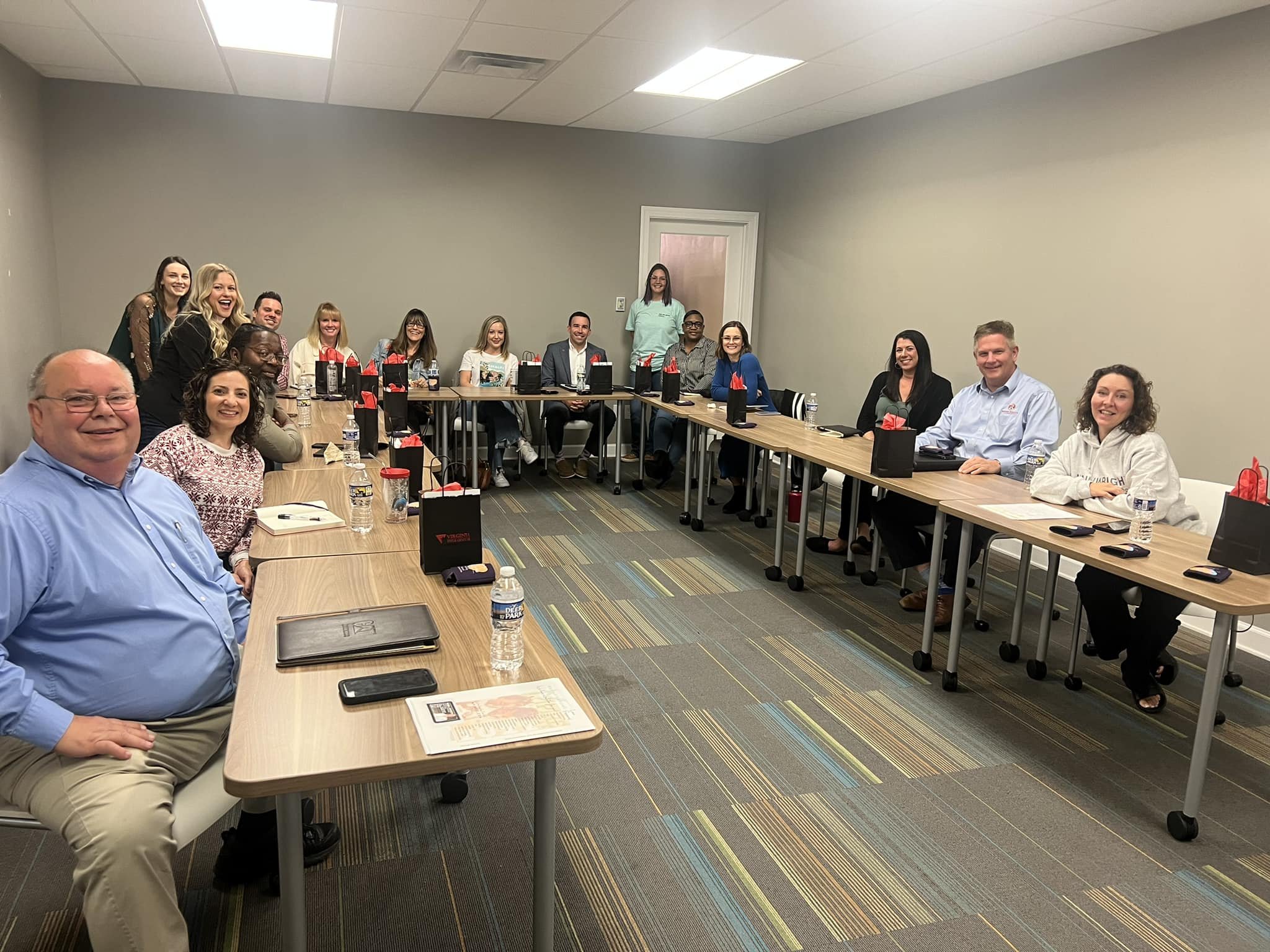 A group of diverse professionals sitting around two long conference tables arranged in a U-shape, smiling for a photo in a meeting room with neutral gray walls and colorful carpet.