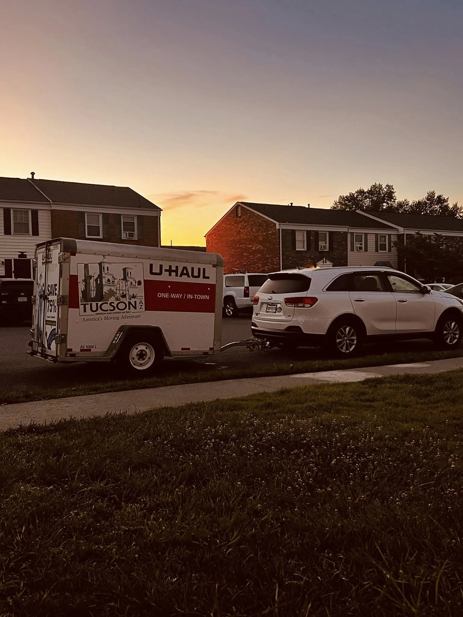 White SUV parked on a residential street at sunset, attached to a U-Haul trailer with houses in the background.