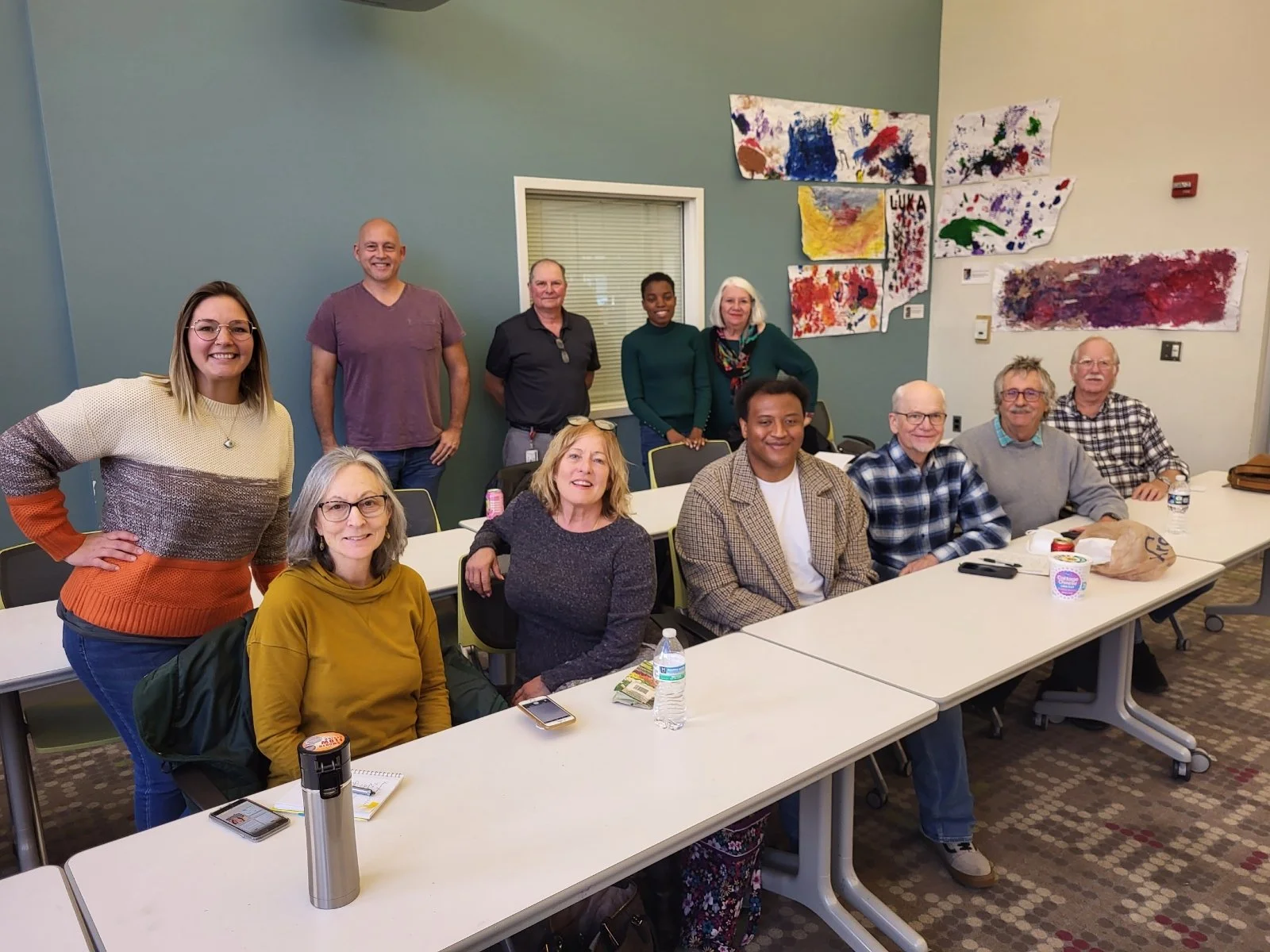 A group of fifteen diverse adults gathered in a classroom or meeting room, smiling for a photo, with colorful artwork on the wall behind them, tables with water bottles, notebooks, and personal items in front of them.