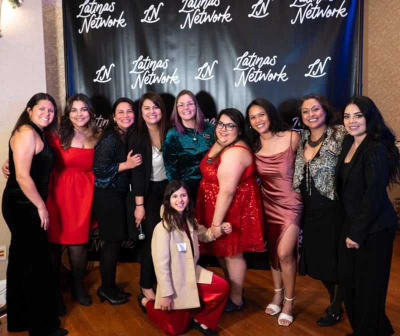 Group of nine women and one young girl posing together in front of a black backdrop with the text "Latinas Network" repeatedly printed on it. The women are dressed in formal and fashionable attire, and they are smiling.