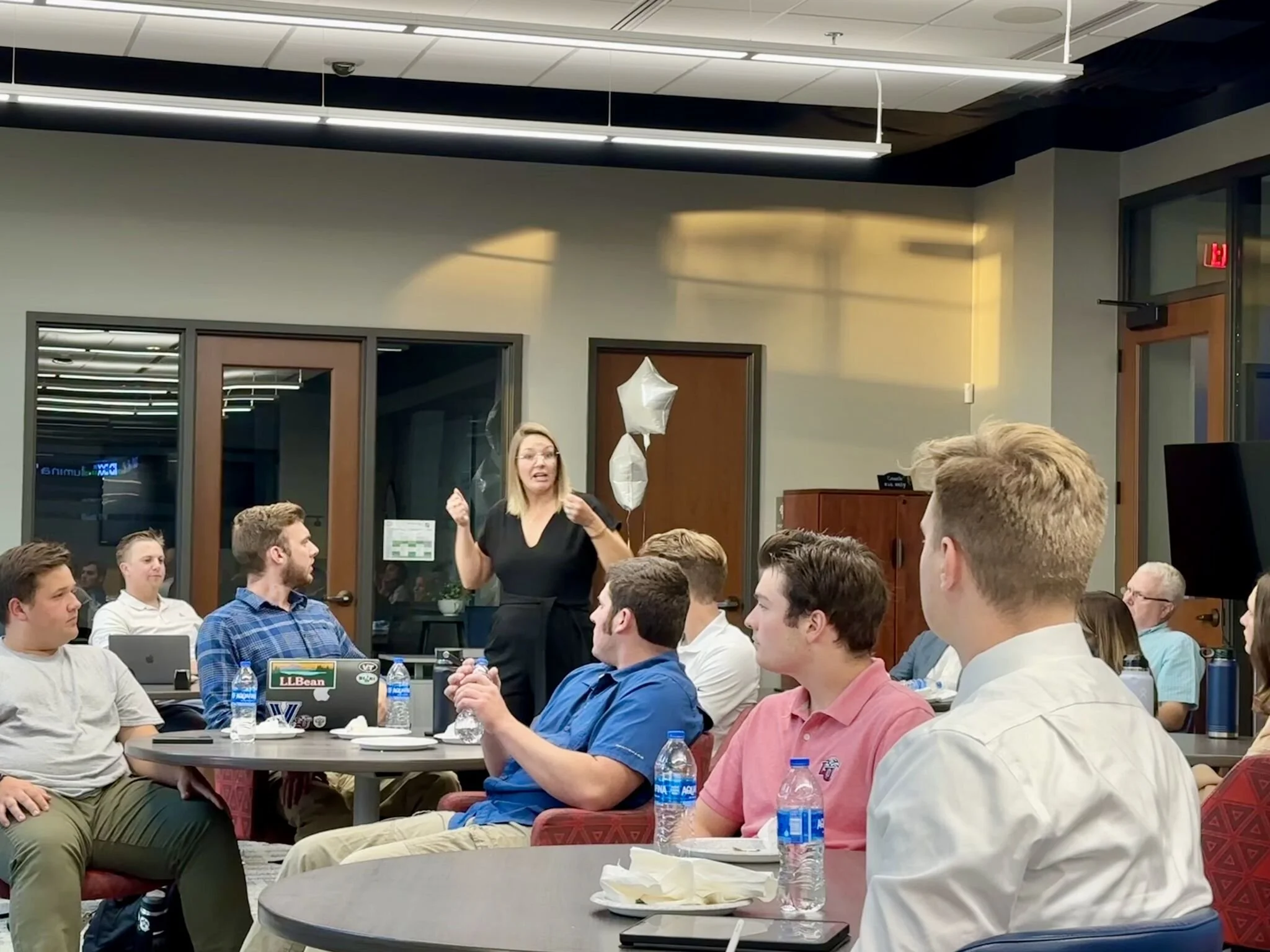 A woman standing and speaking to a group of people seated at round tables in a conference room, with some balloons in the background.