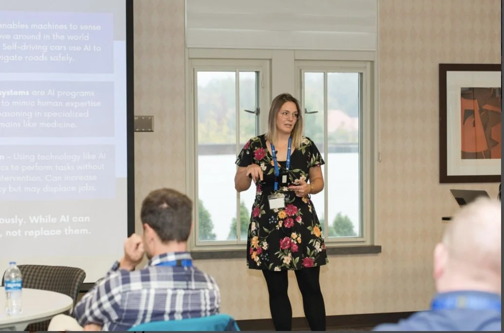 A woman in a floral dress giving a presentation to a group, standing near a window with a projection screen behind her.