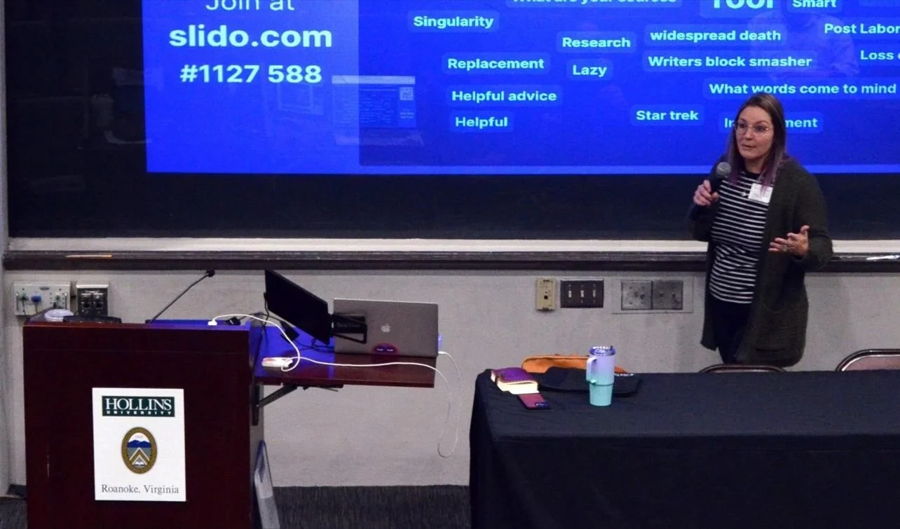 A woman giving a presentation at Hollins University in Roanoke, Virginia, standing next to a large screen with a blue slide showing various words and a URL for Slido.com.
