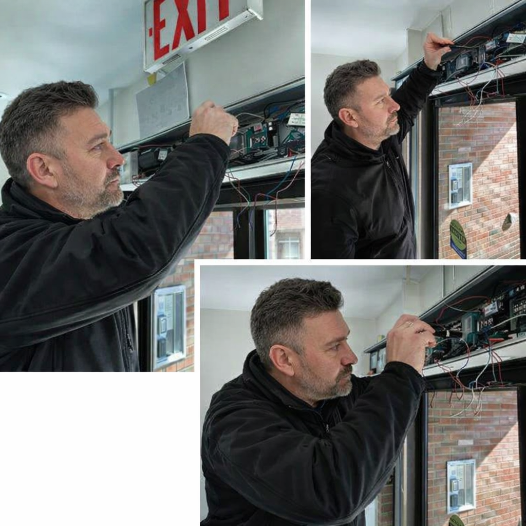 A man working on wiring inside a building near a large window with an exit sign above. The man is handling electrical components or network equipment on a shelf.