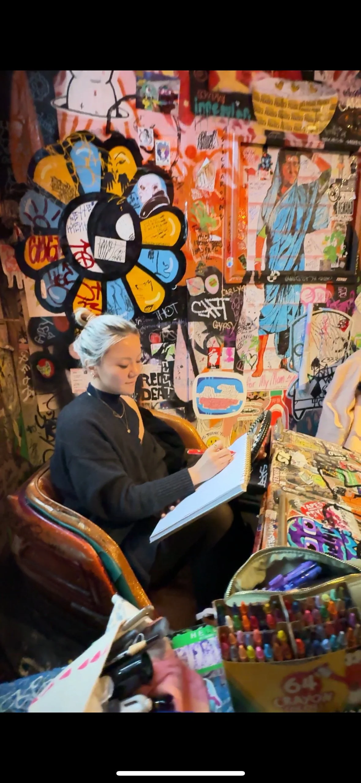 A woman with gray hair, wearing a black hoodie, sitting in a colorful, graffiti-covered space, sketching in a notebook. The wall behind her is filled with vibrant street art and various tags.