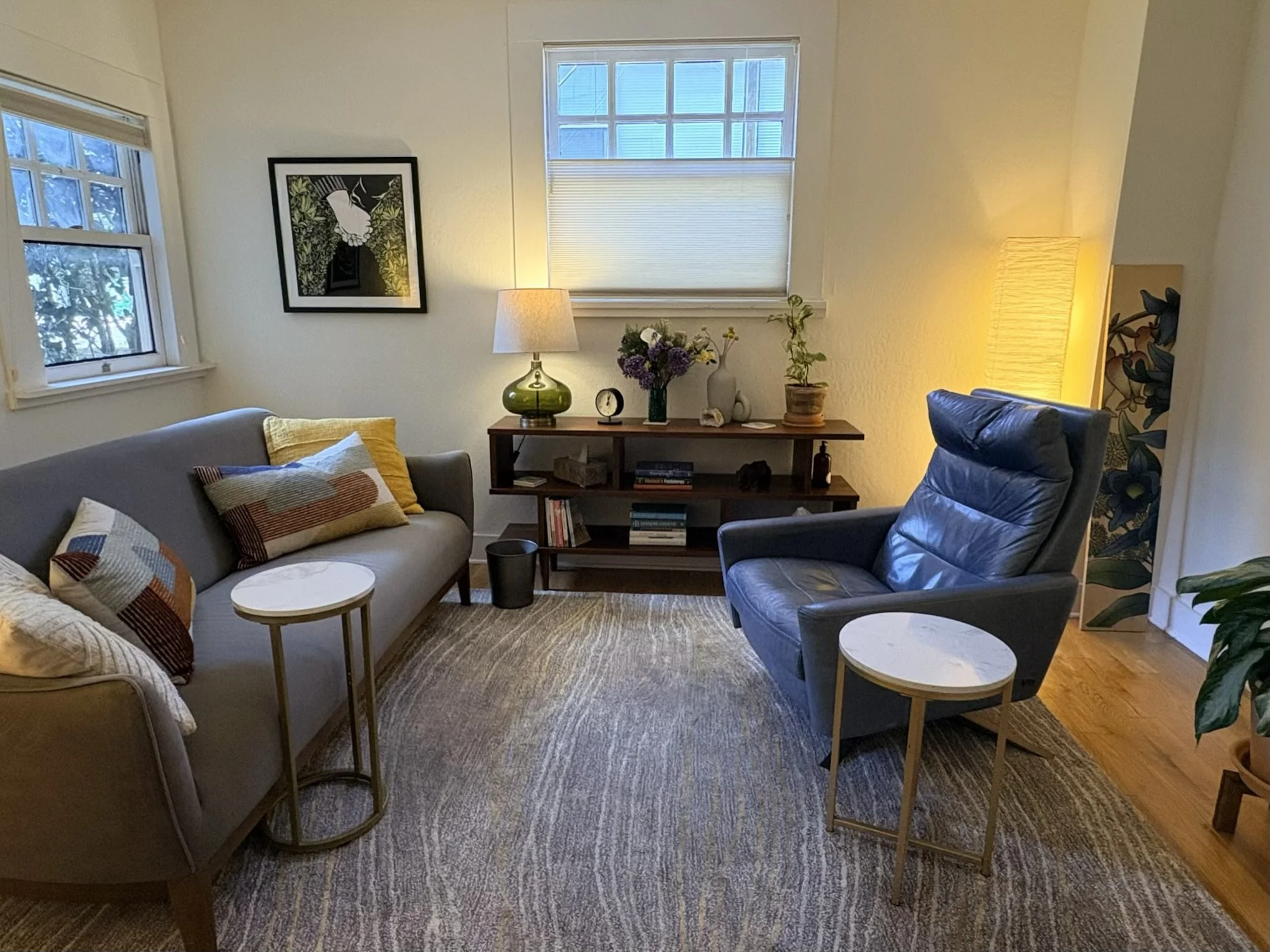 Cozy living room with a gray sofa, navy armchair, and wooden side tables, decorated with colorful pillows, framed artwork, a bookcase with books, and plants near window and bedside lamps