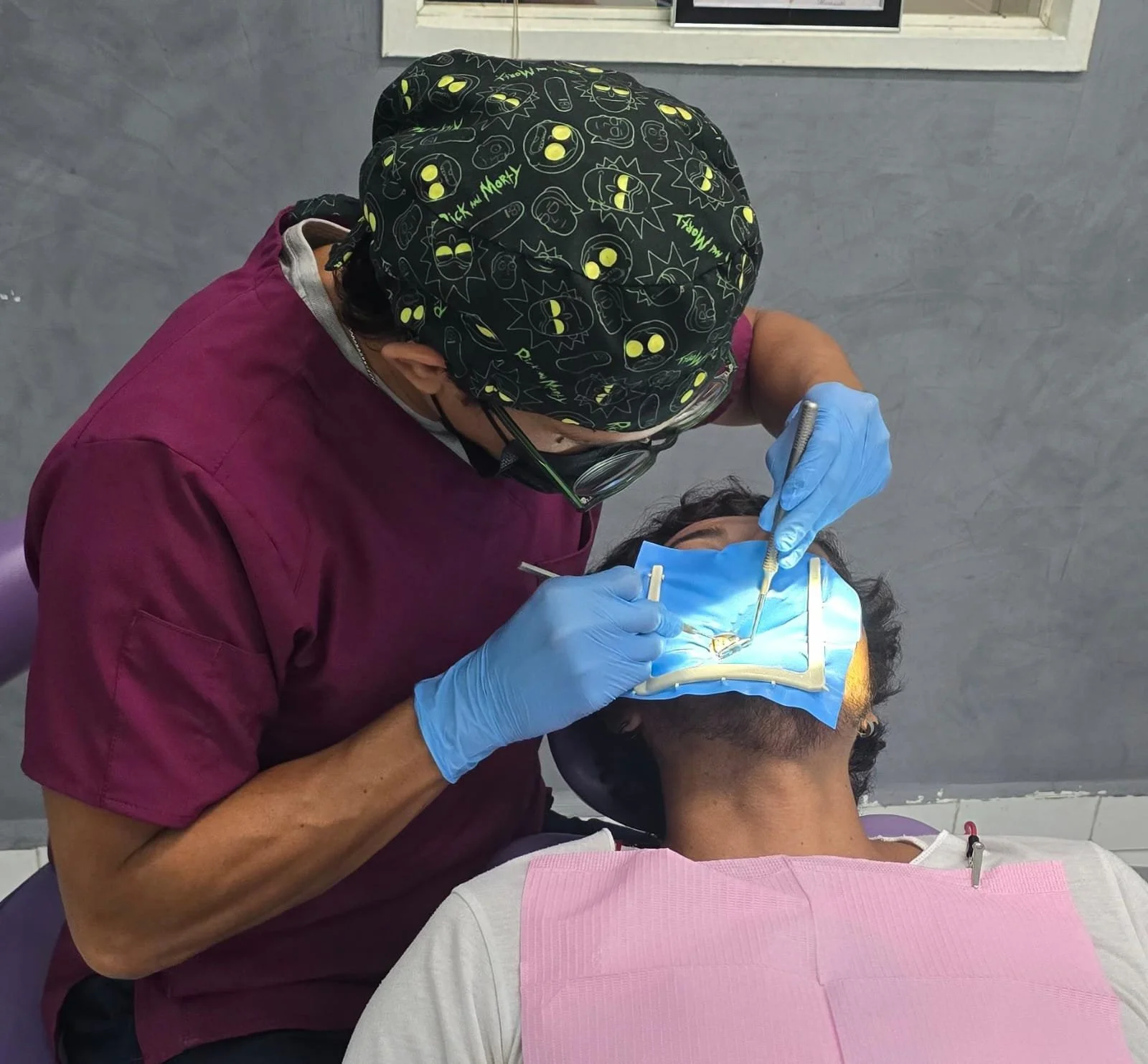 A dentist in purple scrubs and a hat wearing glasses and gloves, working on a patient's mouth in a dental clinic.