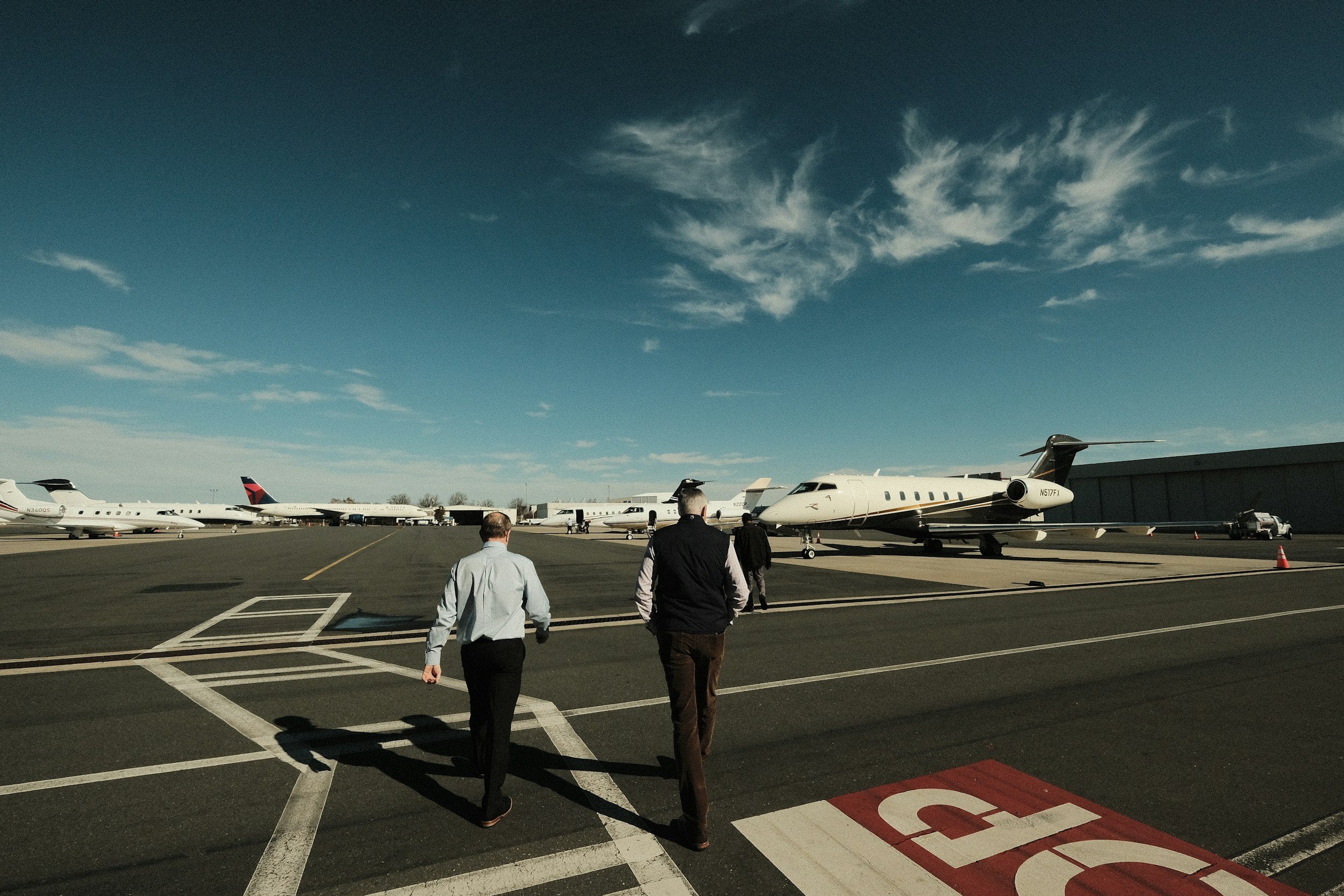 Two men walking on airport tarmac towards private jets parked on the runway under a blue sky with clouds.
