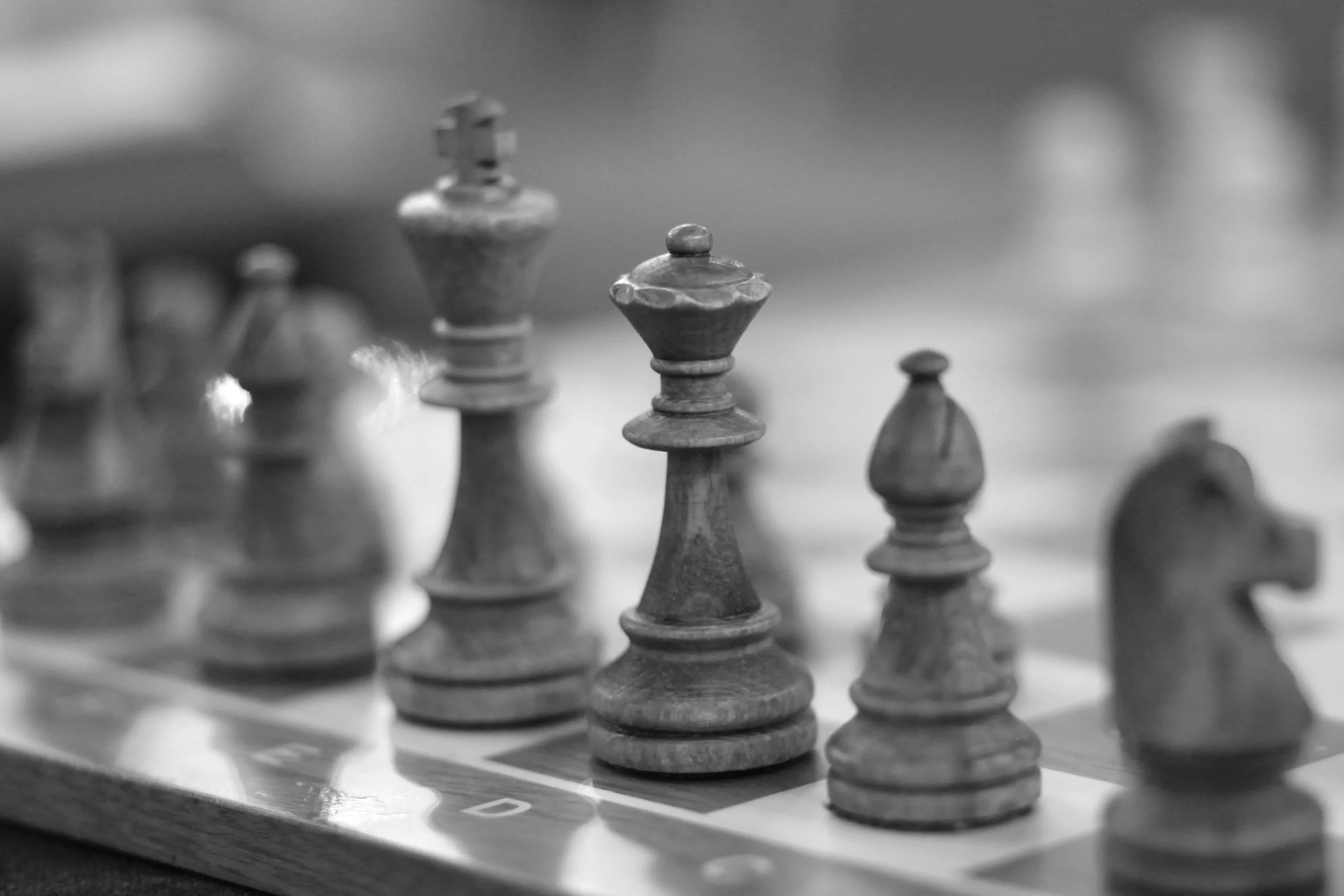 Black and white close-up of chess pieces on a chessboard, with a focus on the queen and king, showing some pieces blurred in the background.