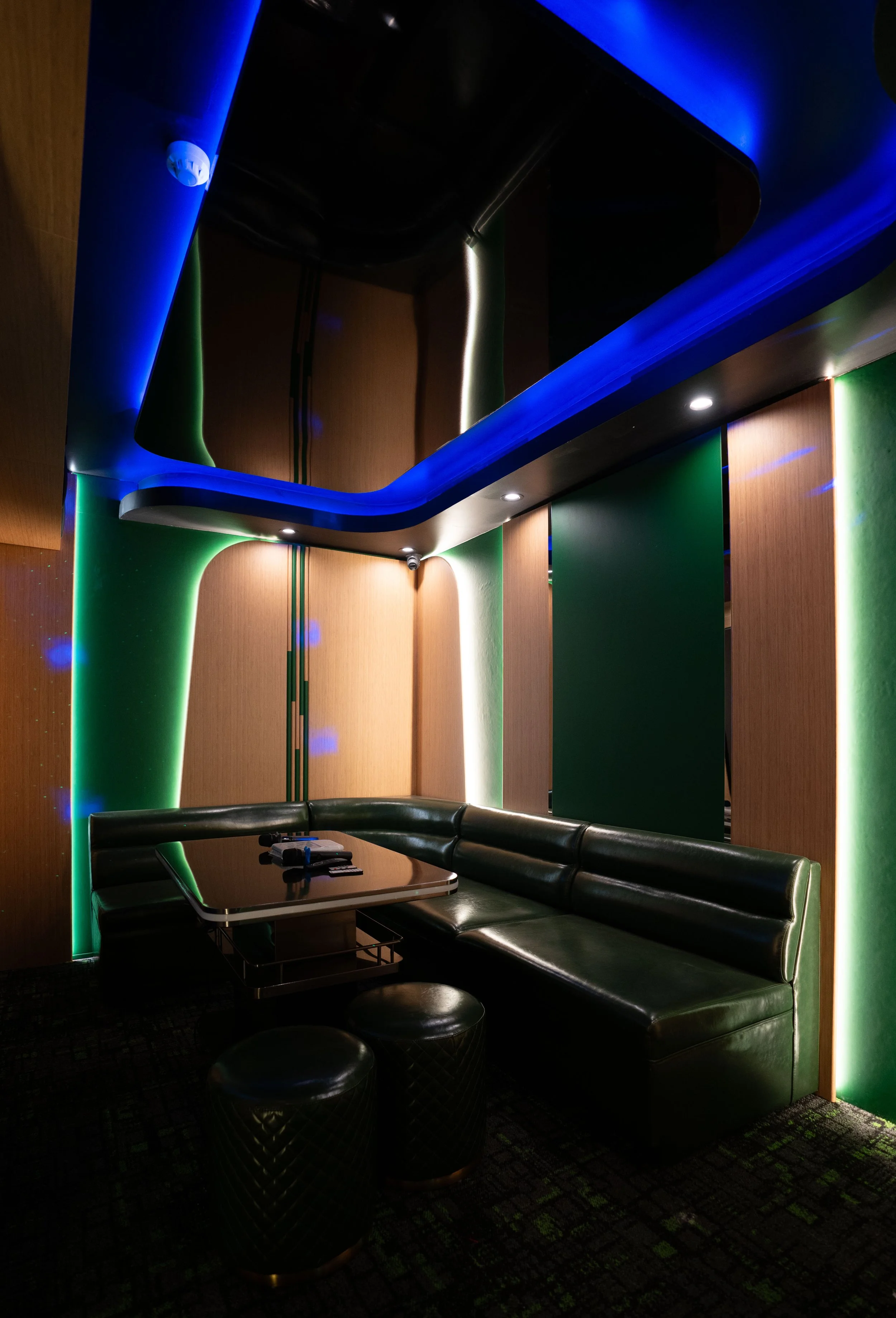A modern lounge area with black leather sectional sofa, small round stools, and a table with remote controls. The walls and ceiling are accented with green, blue, and white lighting, creating a sleek, contemporary ambiance.