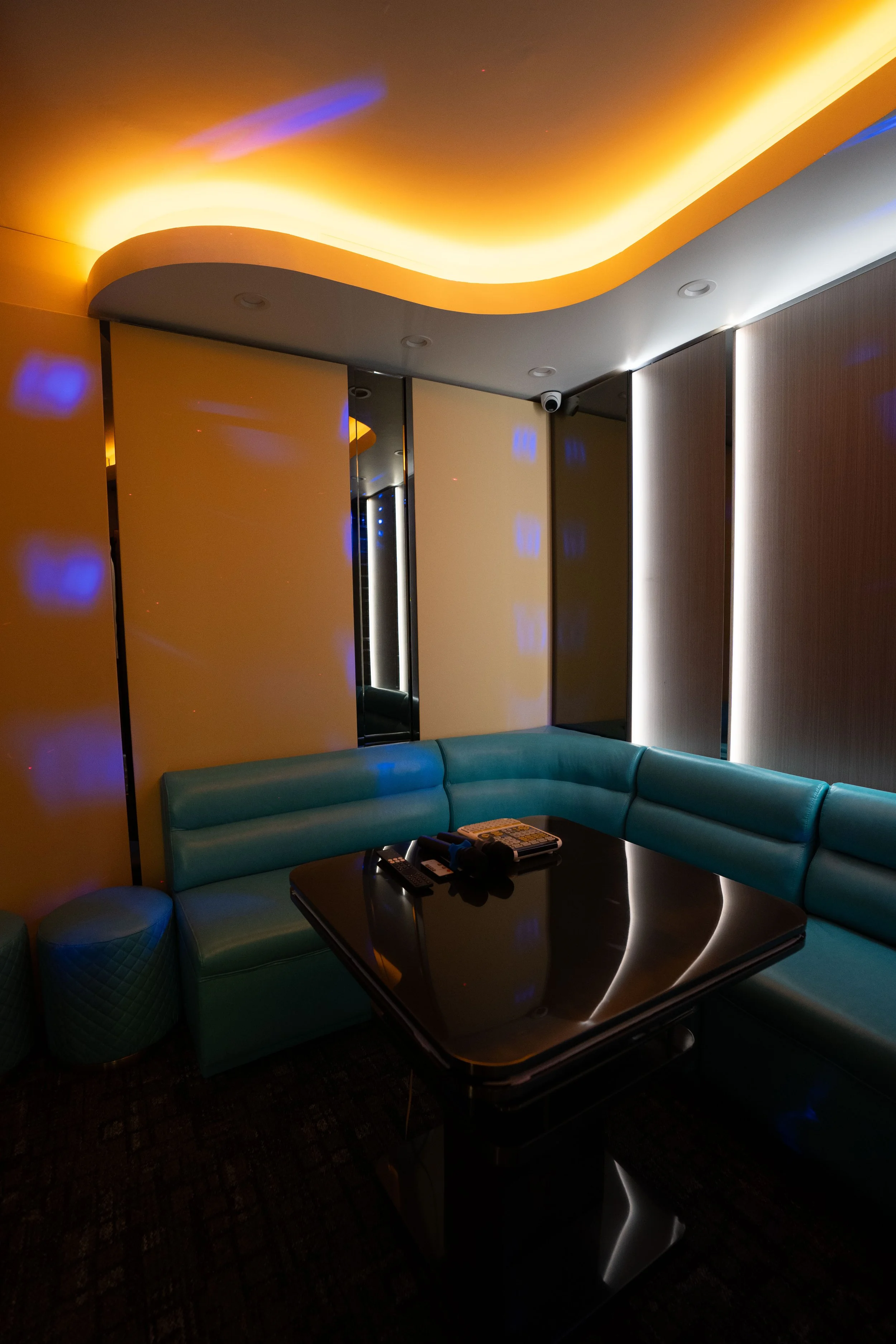 Dimly lit lounge area with curved teal seating, a black coffee table with remote controls and electronic devices, illuminated ceiling with warm yellow and blue lighting, and reflective surfaces.