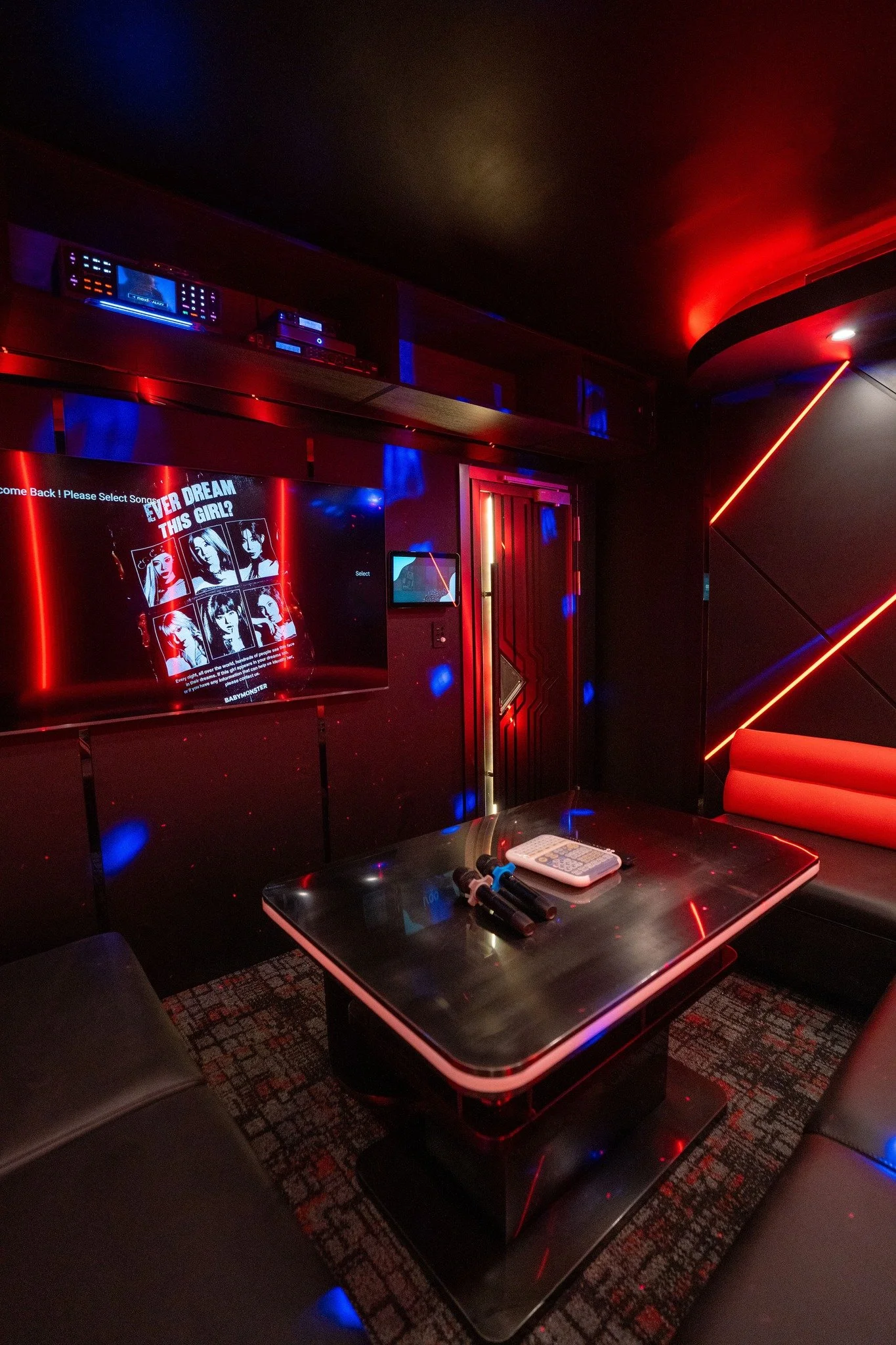 A dimly lit karaoke room with a large screen displaying a song selection with images of women. The room has neon red and blue lighting, a black table with a remote control and microphones, and black leather chairs.
