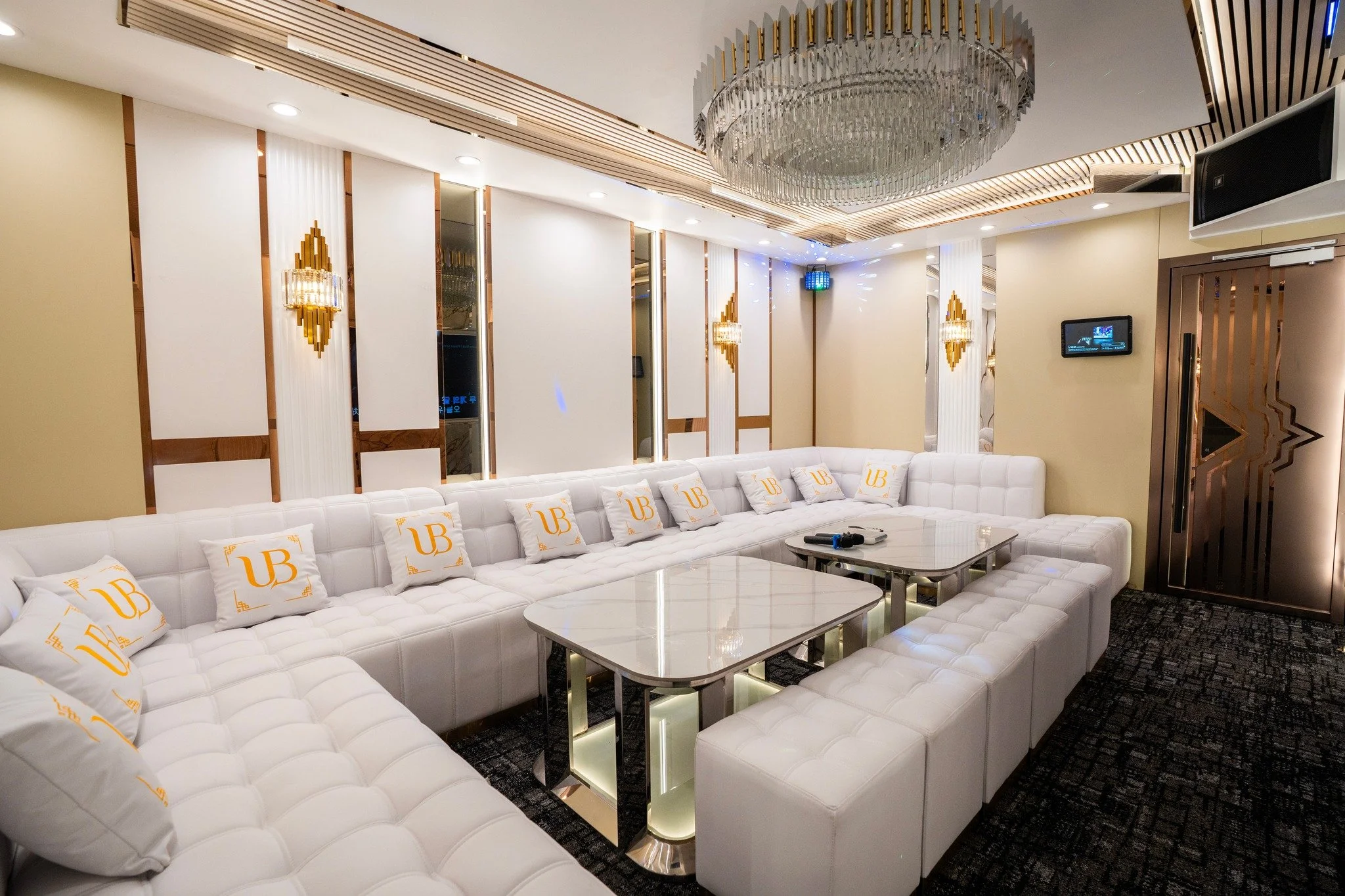 Luxury hotel lobby or lounge with white tufted sofa, gold-accented wall sconces, glass-top coffee tables, and a chandelier on a wood-paneled ceiling.