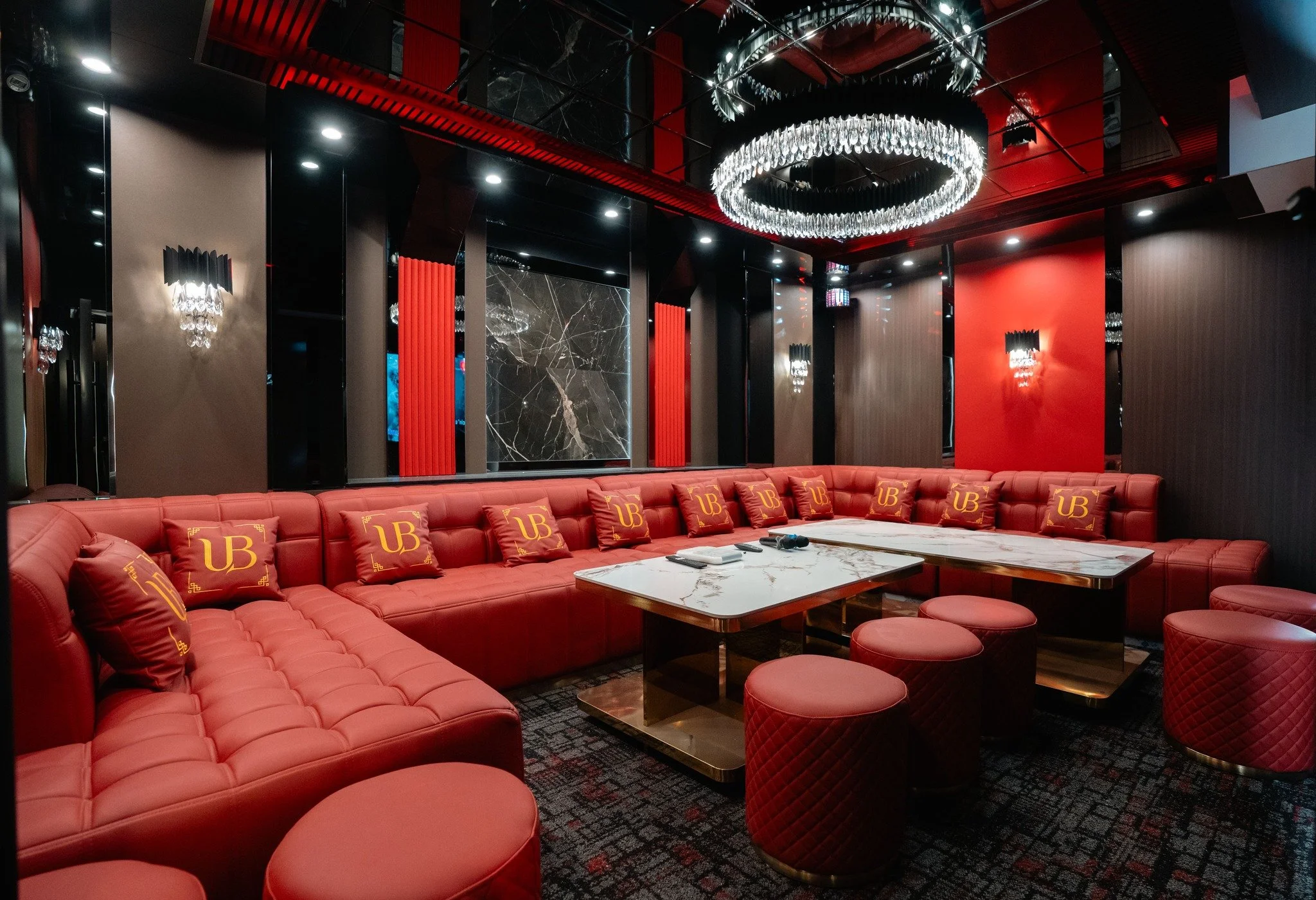 Luxurious lounge with red tufted sofa and scattered pillows, marble-top table, black mirror walls, modern wall sconces, chandeliers, and a dark decor with red and black accents.