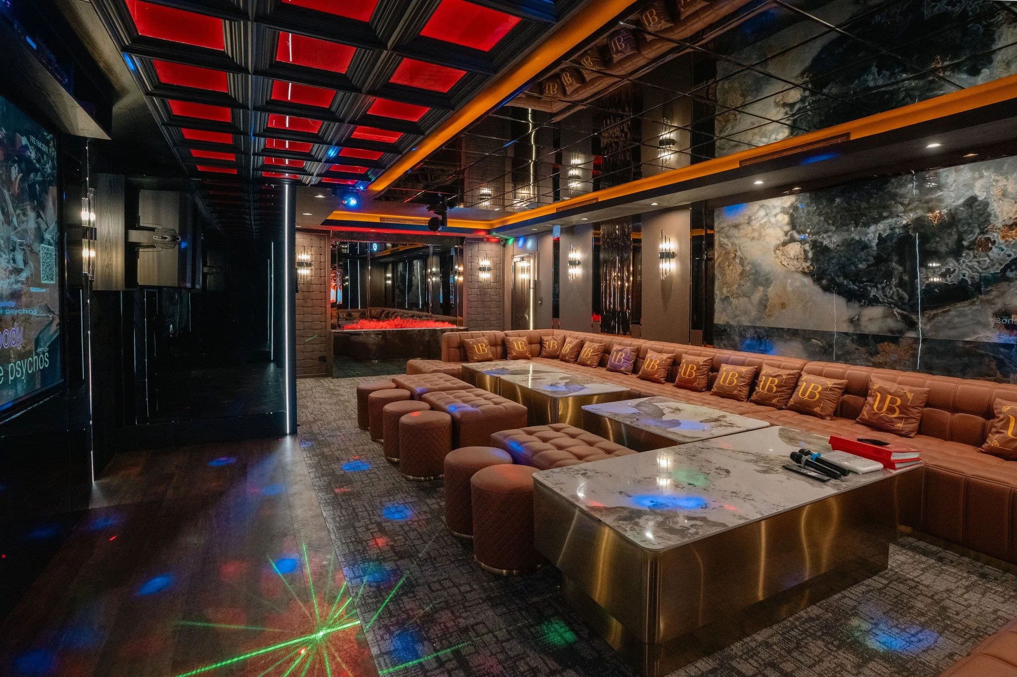 Upscale view of a modern lounge or bar with a black ceiling featuring red panels, a marble table, and tan cushioned seats. The room has dim lighting and colorful LED accent lights.